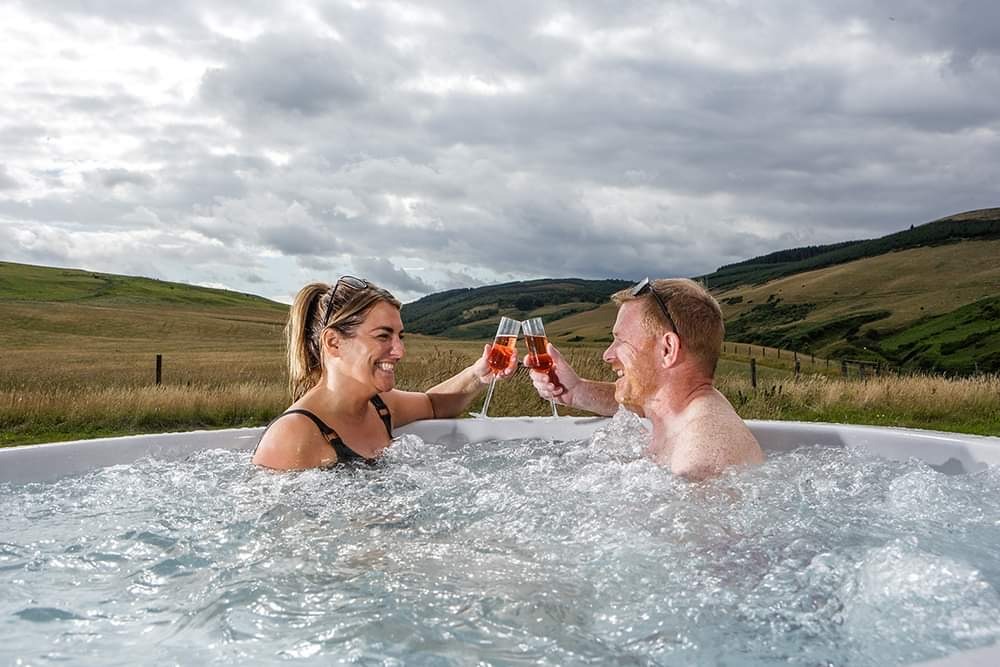 A man and a woman are sitting in an outdoor hot tub at Pathgreen, toasting with drinks in their hands. Surrounded by rolling green hills under a cloudy sky, they are smiling and look relaxed, enjoying the scenic landscape and the warm water during their glamping getaway.