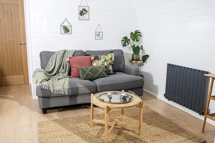 A cozy Pathgreen living room with a gray couch adorned with green and pink pillows and a green throw. A round wooden coffee table sits on a woven rug. Potted plants and framed botanical prints decorate the space, bringing a touch of glamping indoors. A wooden door and black radiator are in the background.