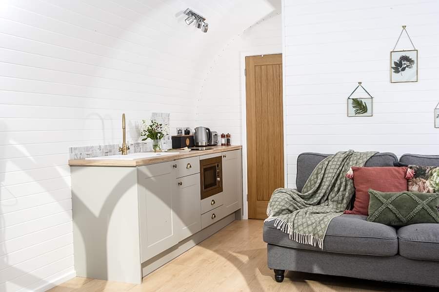 A small, modern kitchen with light wood countertops and white cabinets. It features a sink, stove, and various appliances. Adjacent to the kitchen is a cozy living area with a gray sofa, green and red cushions, and a green throw blanket from Pathgreen. The walls are white paneled.