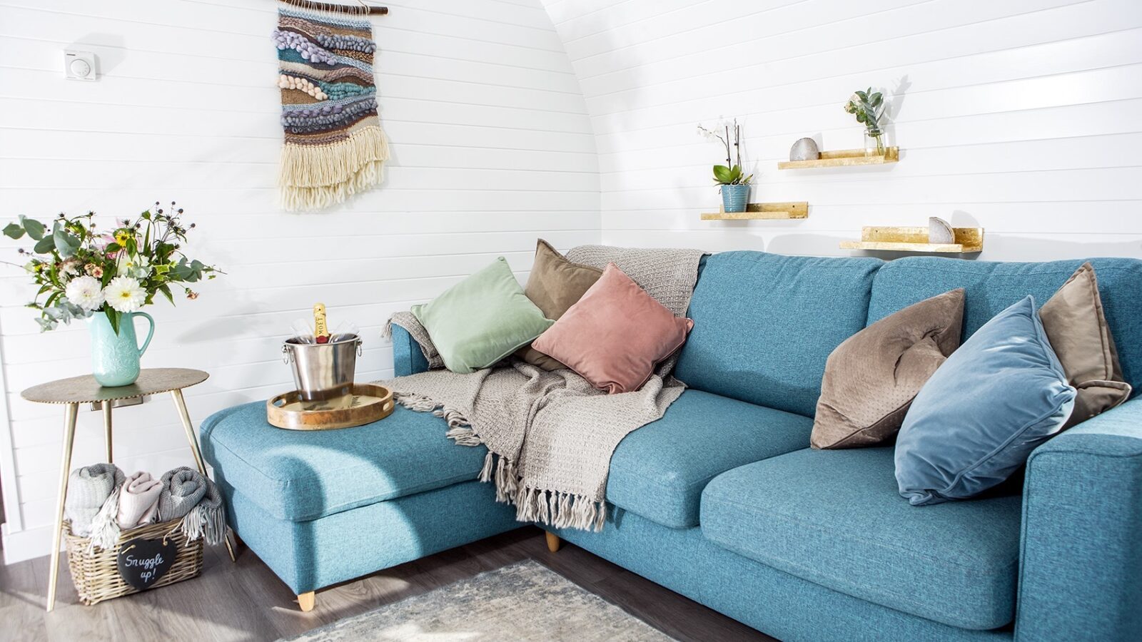 A cozy living room featuring a blue sectional sofa adorned with various pillows and a gray throw, reminiscent of glamping comfort. On the side table, a bouquet of flowers sits in a teal jug alongside a bucket of champagne. A woven wall hanging decorates the white shiplap wall.