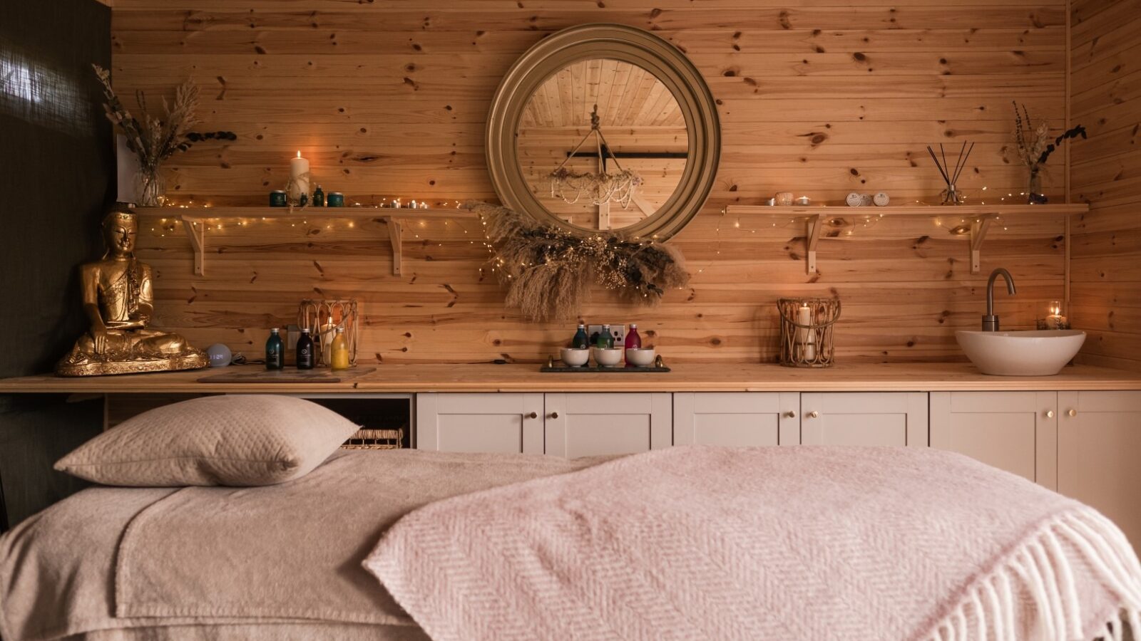 A cozy spa room with wooden walls and ceiling. A massage table covered with a soft pink blanket and pillow is centered. Shelves holding candles, oils, and decor line the back wall. A round mirror and a golden statue add elegance and tranquility to this eco-friendly haven.