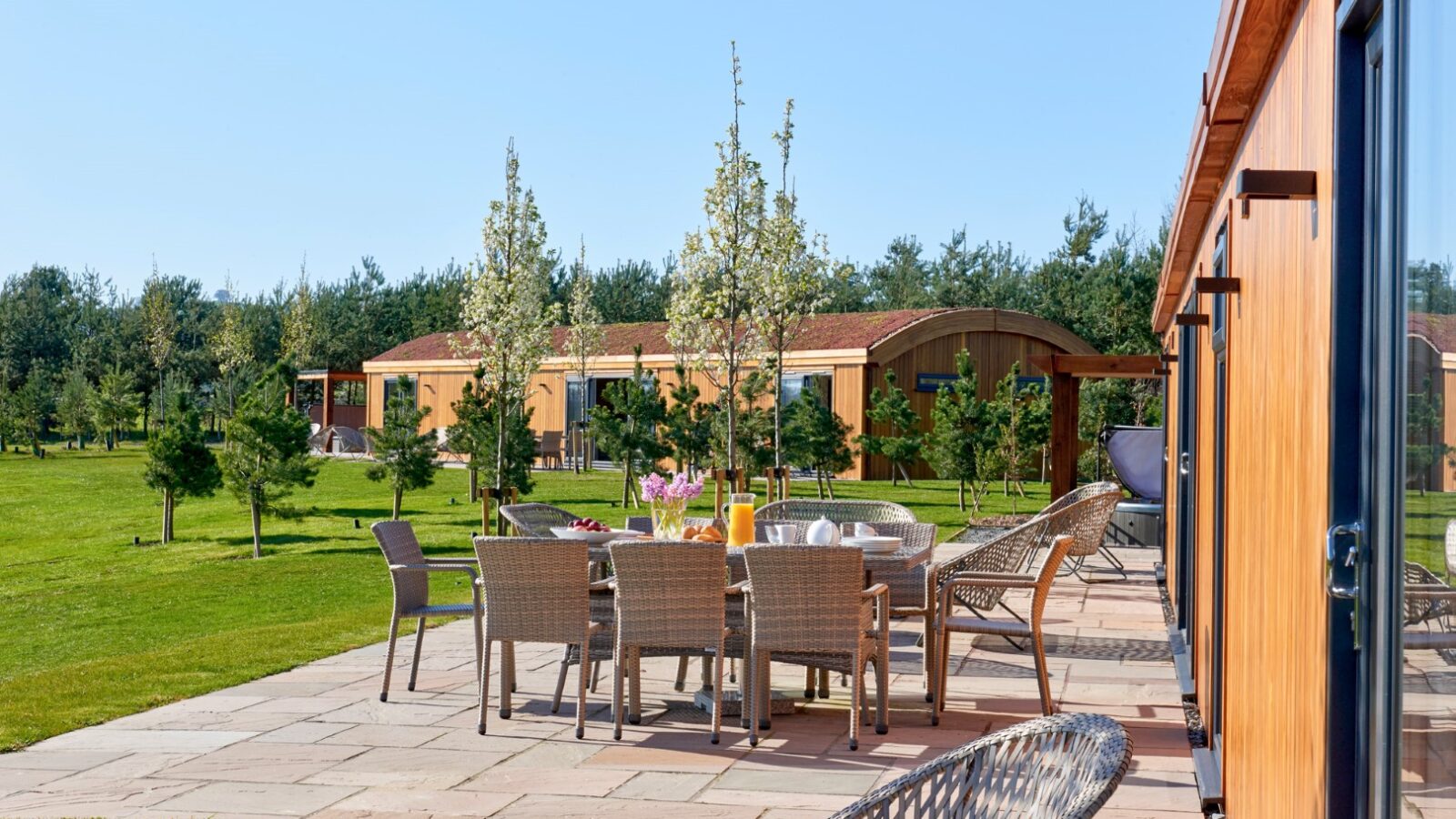 An outdoor patio with a dining set and lounge chairs, flanked by a wooden building on a neatly paved surface. The background displays lush parkland with green lawns, several young trees, and other similar cabin-like lodges under bright, clear skies.