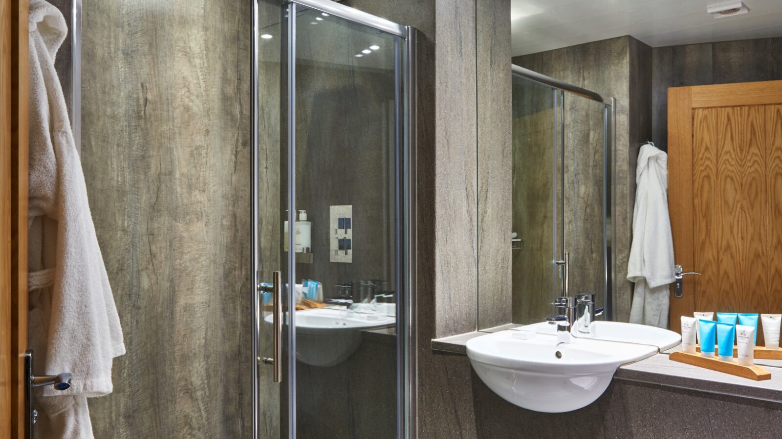 A modern bathroom in Parkland Lodges features a glass-enclosed shower, wooden accents, and a counter with two sinks. Blue toiletries and white towels are neatly arranged on the counter. A wooden door and a white bathrobe hanging on a hook add to the serene ambiance.