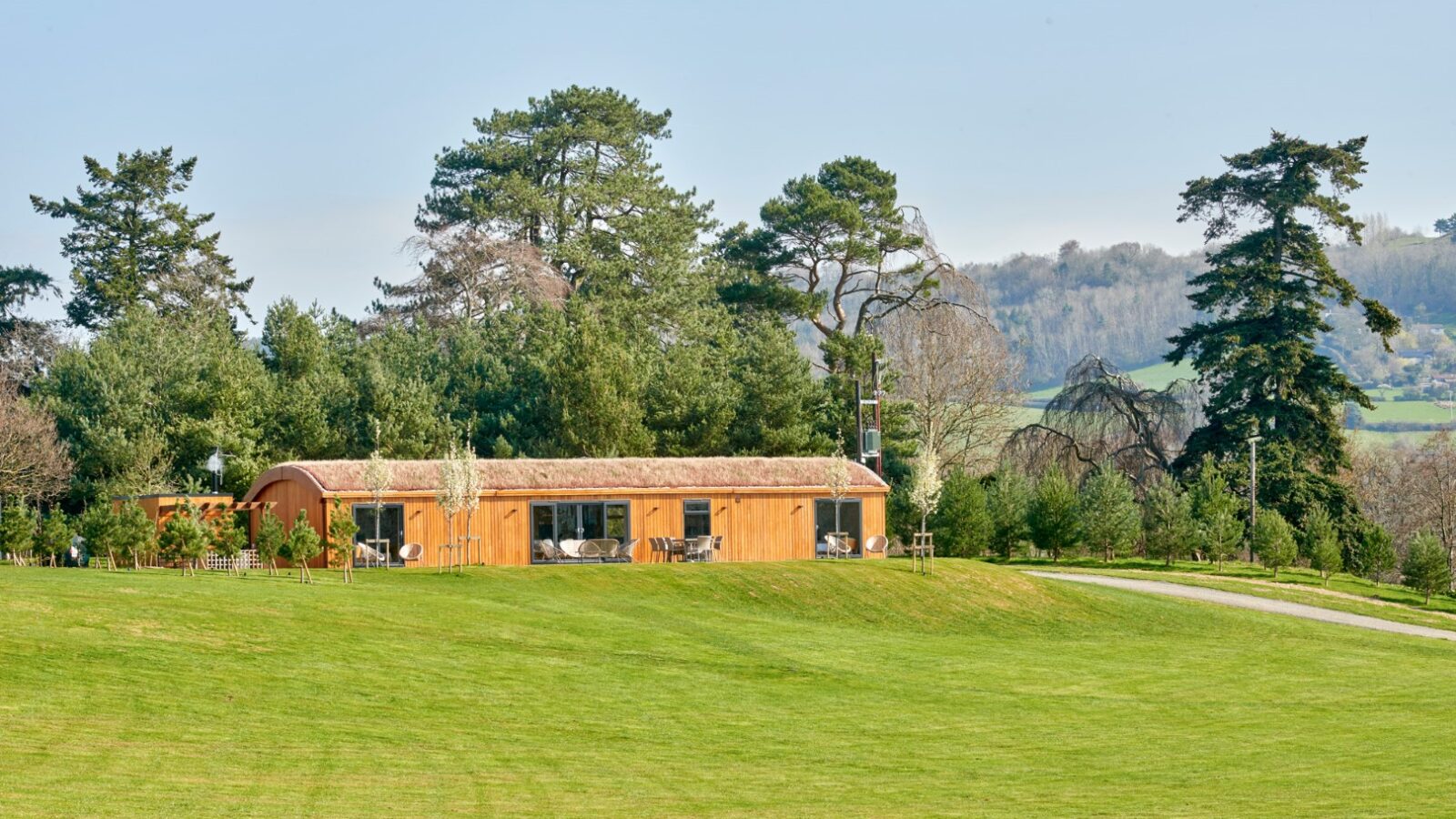 A modern, single-story wooden lodge with large windows, surrounded by tall trees and greenery, is situated in scenic parkland. The grassy field in the foreground slopes gently upward towards the structure, with distant hills visible in the background.