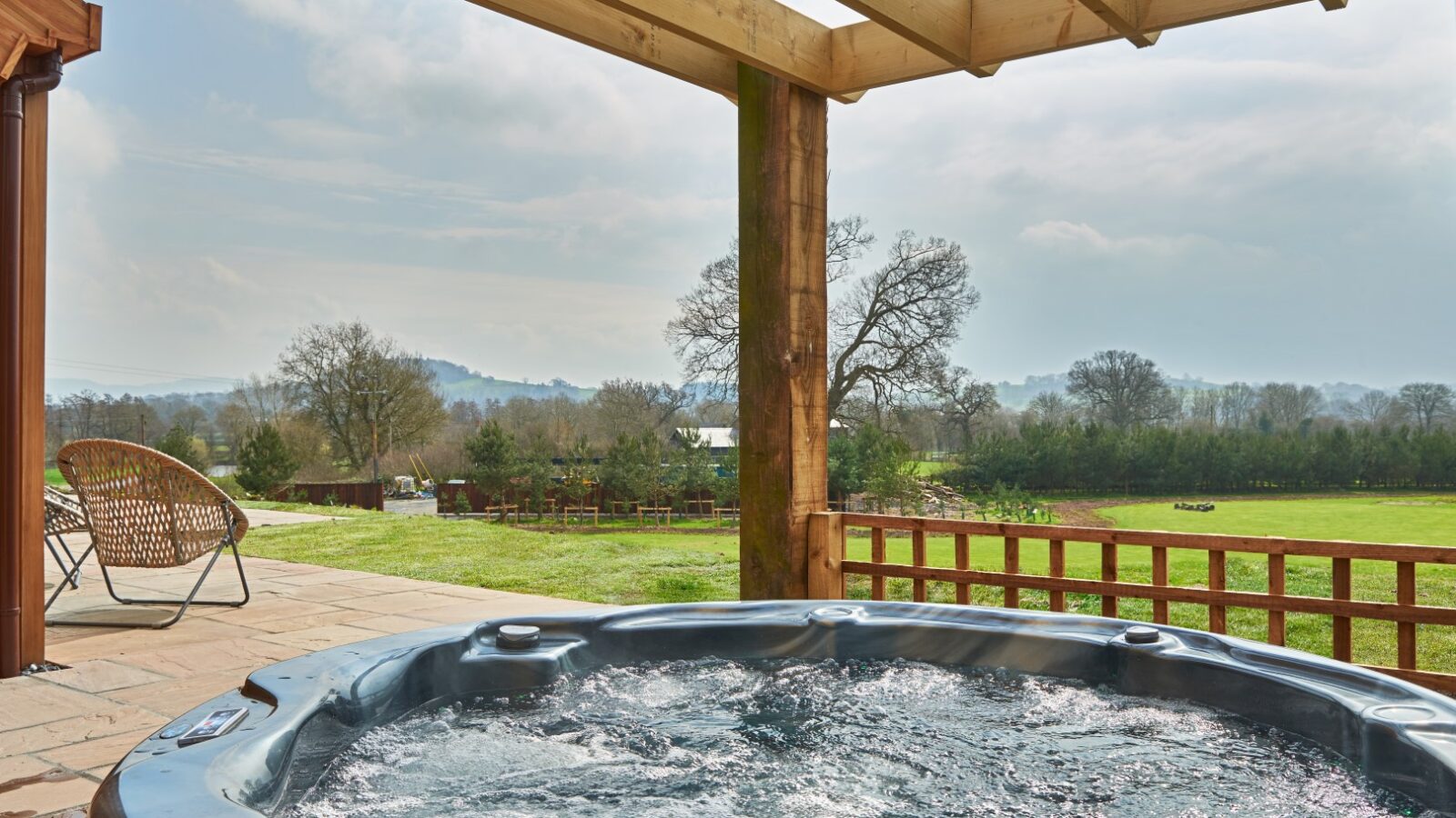 A hot tub with bubbling water is situated on a patio with wooden pillars and an overhead pergola. In the background, there is a vast green field, parkland trees, and a distant view of a hilly landscape under a cloudy sky. A wicker chair is on the left side of the patio.