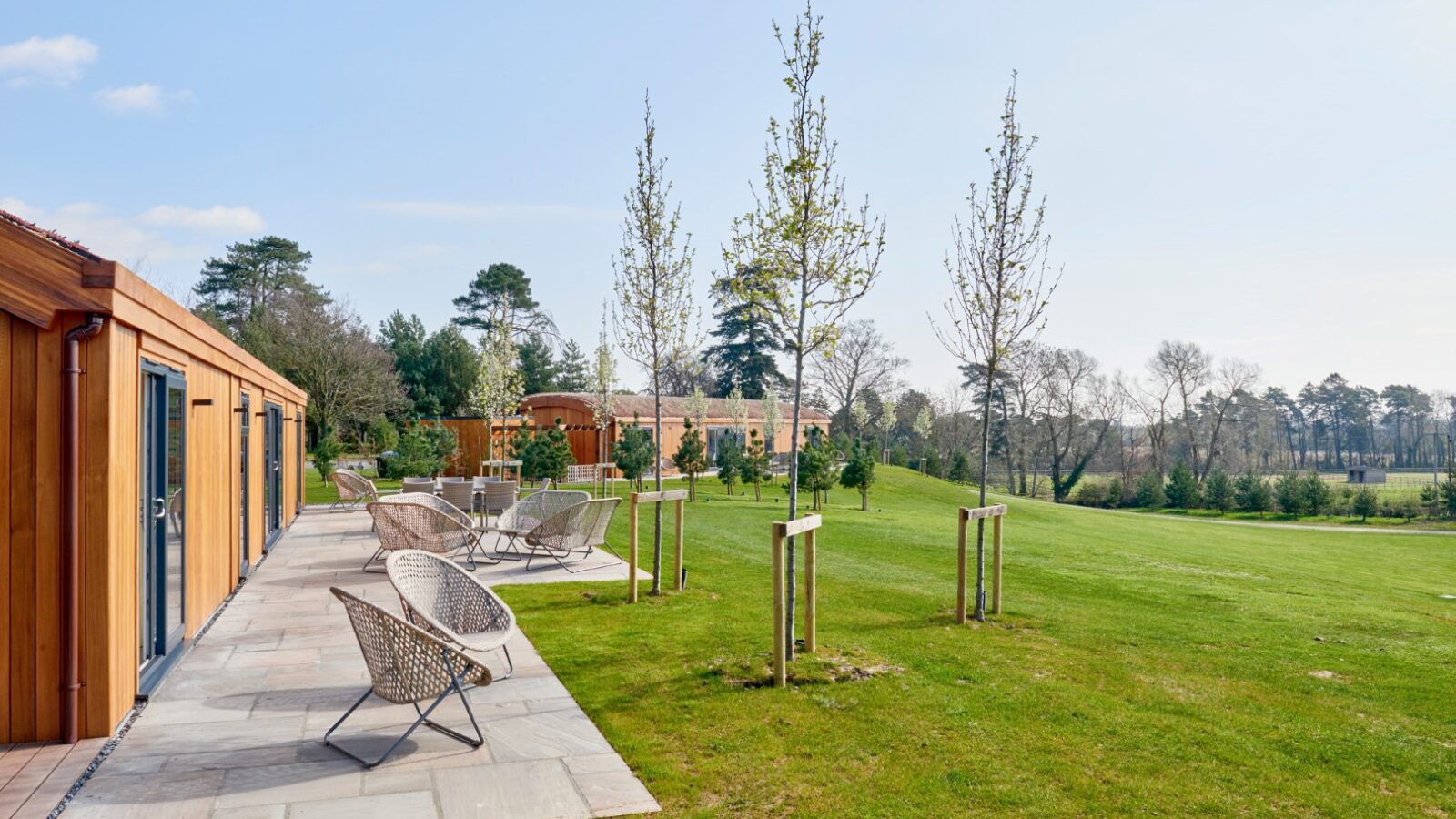 A spacious outdoor patio with wicker chairs and tables overlooks a green lawn with young trees. Modern lodges with wooden siding are visible to the left, surrounded by lush foliage and parkland under a clear blue sky in the background.