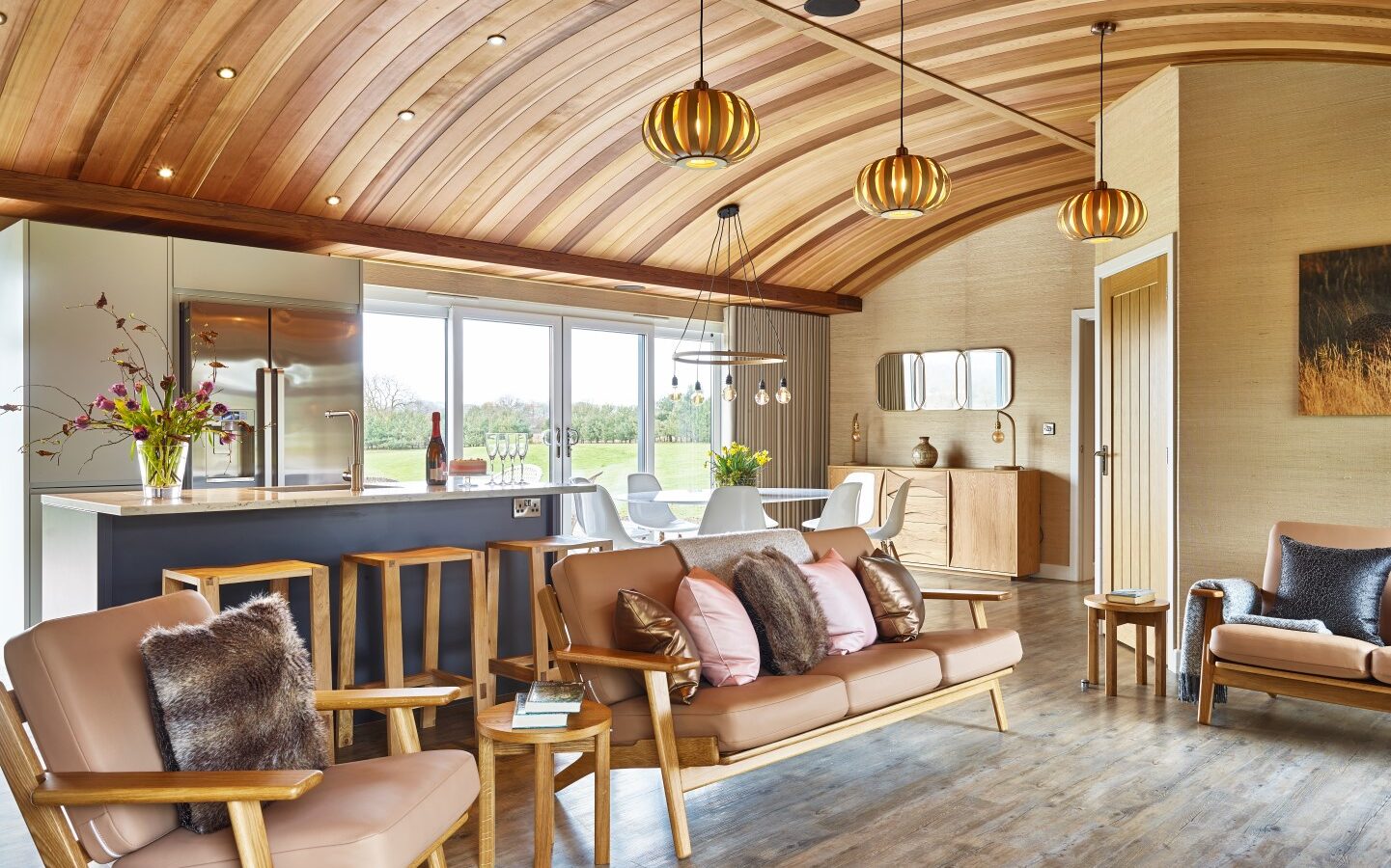 A spacious, modern living area with a curved wooden ceiling and ample natural light. It features a kitchen and dining area with a stainless steel fridge, contemporary light fixtures, and seating arrangements with brown leather sofas adorned with cushions and throws—perfect for lodges surrounded by parkland.