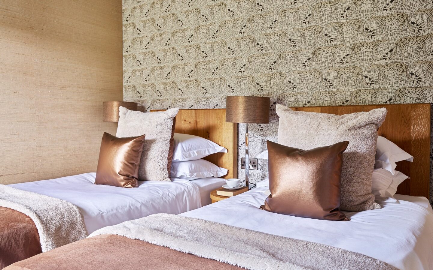 A warmly lit bedroom reminiscent of cozy lodges, with two single beds adorned in white linens, brown throws, and matching brown and beige pillows. Between the beds is a small wooden nightstand with a brown lamp. The accent wall has patterned wallpaper featuring cheetahs, perfect for nature lovers in parkland settings.