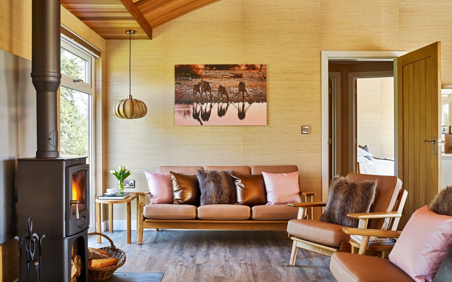 A cozy living room reminiscent of charming lodges, featuring a modern wood-burning stove, a wicker basket of logs, two tan leather sofas with pink and brown pillows, a wooden coffee table with a potted plant, and a framed horse photo on the wall. The room features wooden flooring and a curved wooden ceiling.