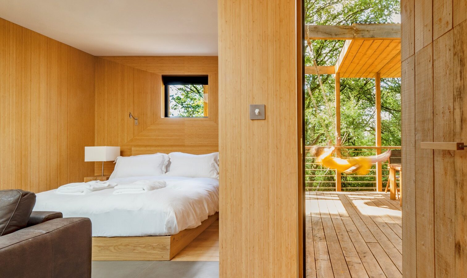 A minimalist bedroom with wooden walls and flooring features a white bed, a small window, and a side table with a lamp. Through an open door, a wooden deck with a yellow hammock is visible, surrounded by green trees and natural light, giving the space the cozy feel of a treehouse.