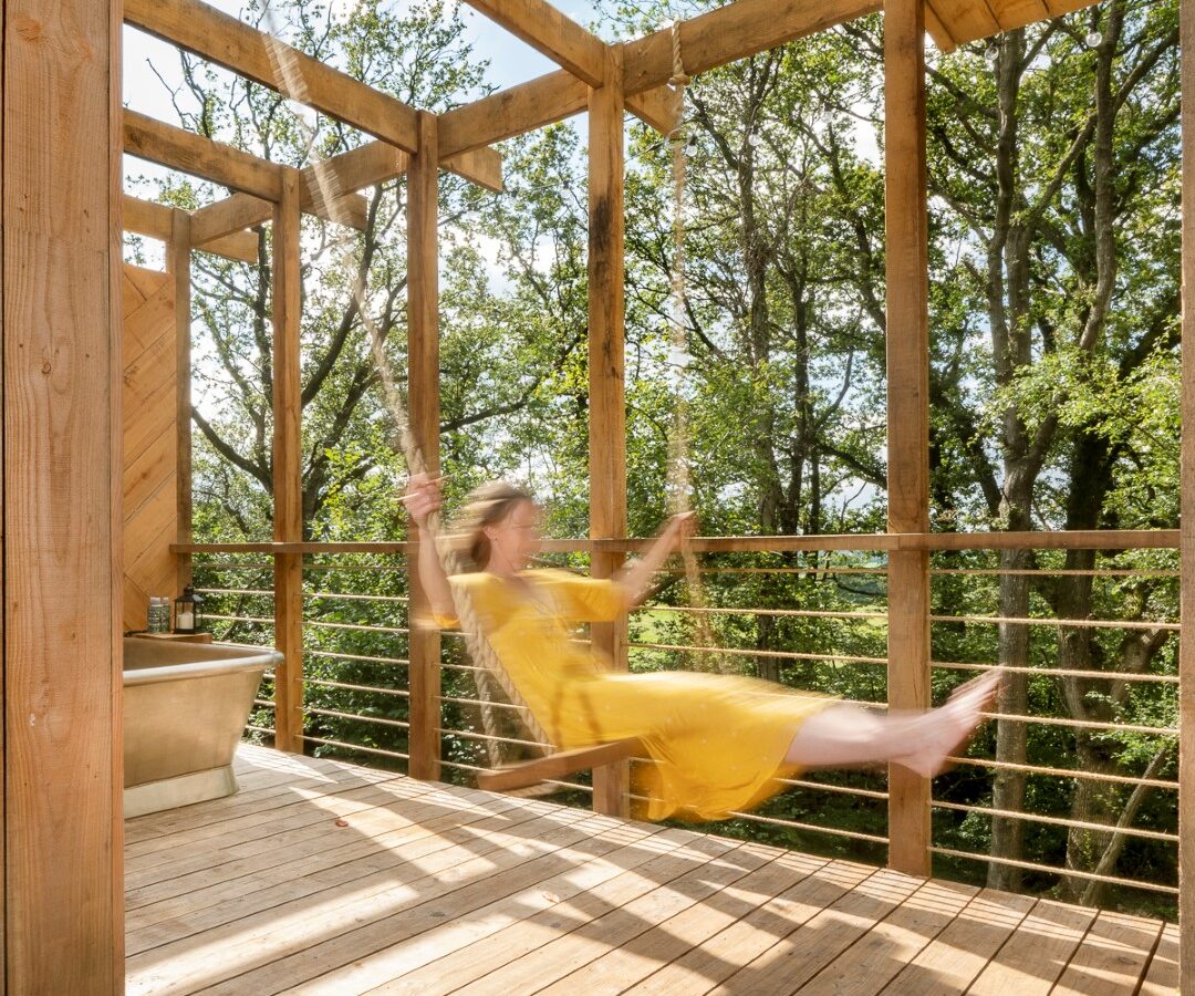 A person in a yellow dress enjoys swinging on a wooden swing set on a spacious, sunlit wooden deck surrounded by trees. The swing is attached to the beams of the deck, resembling a whimsical treehouse setting, and the person appears to be in mid-swing, captured in a motion blur.