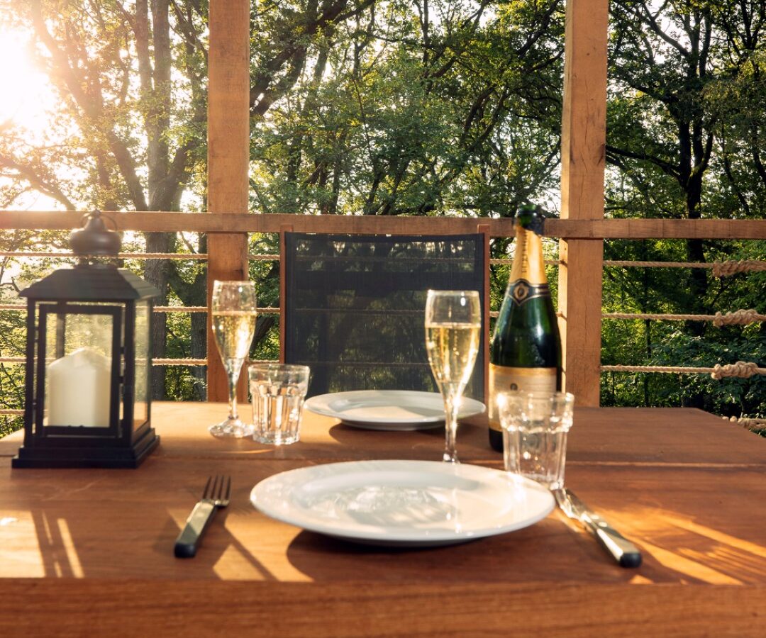 A wooden table set for two with plates, glasses, and utensils, positioned on a sunlit deck of a charming treehouse overlooking lush green trees. A lantern, pinwheel, bottle of champagne, and filled glasses add a cozy and romantic touch as sunlight filters through the trees creating a warm ambiance.