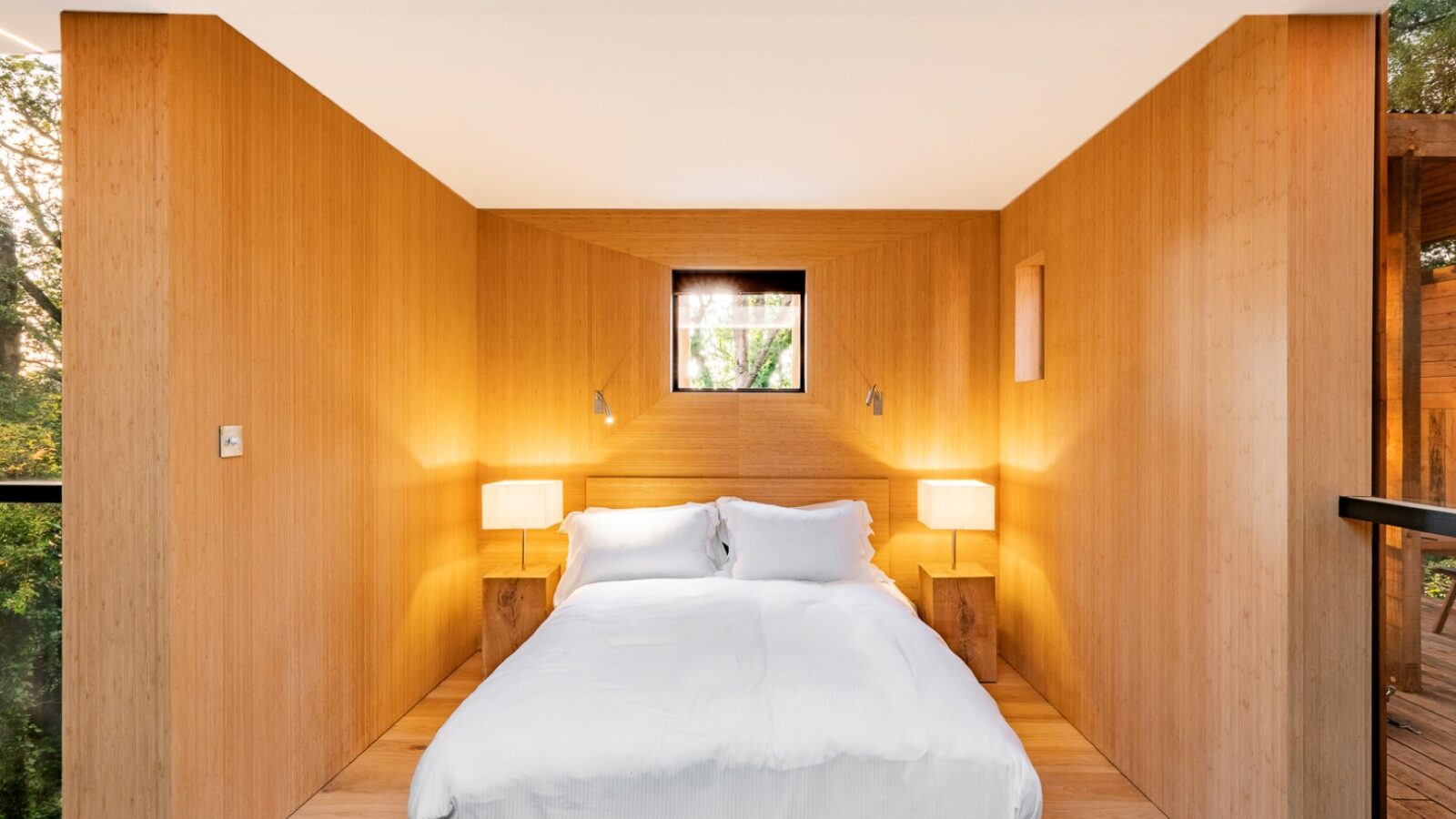 A minimalist bedroom with wooden walls evokes the charm of a cozy treehouse. A large bed with white bedding is centrally placed, flanked by two small bedside tables each with a lamp. A window allows natural light to enter, and greenery visible outside adds to the inviting decor of this treehouse rental.