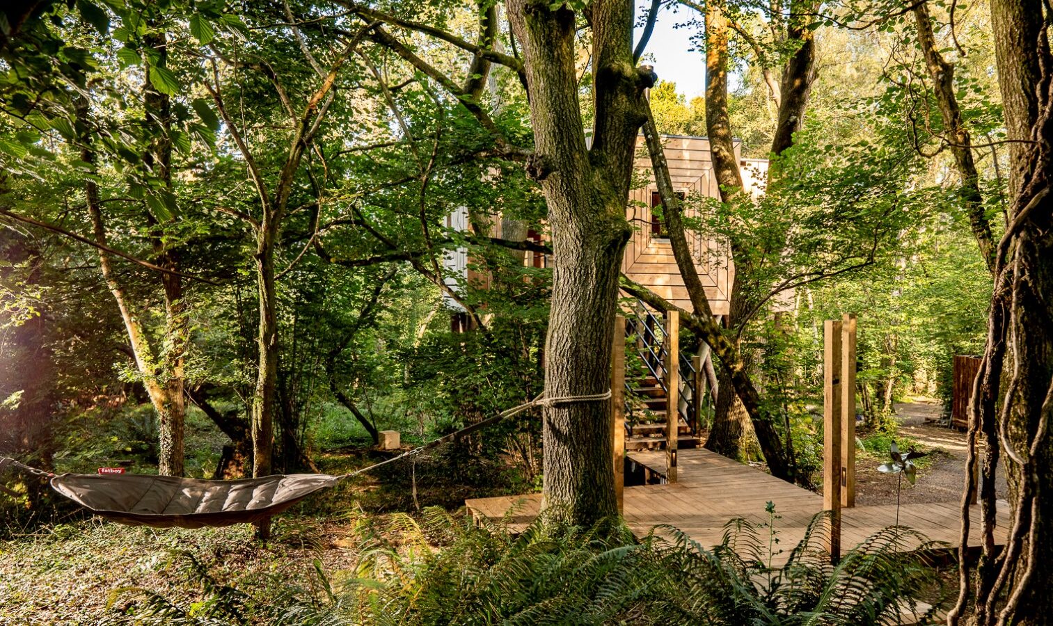 A serene wooded area featuring a wooden treehouse partially obscured by trees. A hammock hangs between two trees in the foreground, and a wooden pathway leads up to the treehouse, where a colorful pinwheel spins gently in the soft, natural sunlight filtering through the leaves.