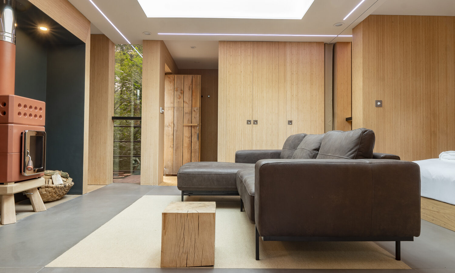 A modern, cozy living space named the Pinwheel Treehouse features a brown sectional sofa, a wooden cube coffee table on a light rug, and a wood-paneled accent wall. A unique red wood-burning stove stands in the corner next to stacked firewood. Large window views of greenery provide natural light.