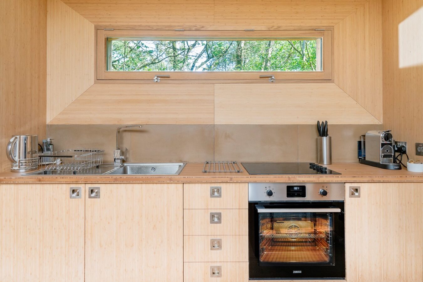 A modern kitchen with wooden cabinetry, featuring a stainless steel sink, electric oven, stovetop, and countertop appliances like a coffee maker and kettle. Above the counter, there's a landscape window offering a view of green trees outside, reminiscent of a serene treehouse rental.