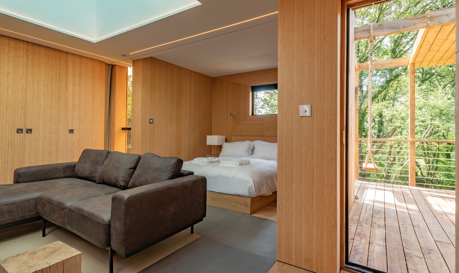 Modern, minimalist bedroom with wooden walls and floors, reminiscent of a cozy treehouse. A large window illuminates the space, enhancing the warm tones. A grey L-shaped sofa faces a low wooden table. A queen-sized bed with white linens sits near another window. An open door leads to a wooden deck outside.