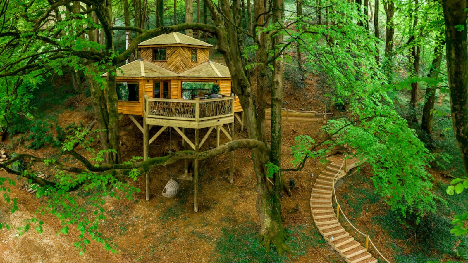 The Ravendere Treehouse sits elevated among lush green trees in a forest. It features two hexagonal sections with shingled roofs and a large wraparound balcony. A winding wooden walkway leads up to the entrance, integrated seamlessly with the natural surroundings.