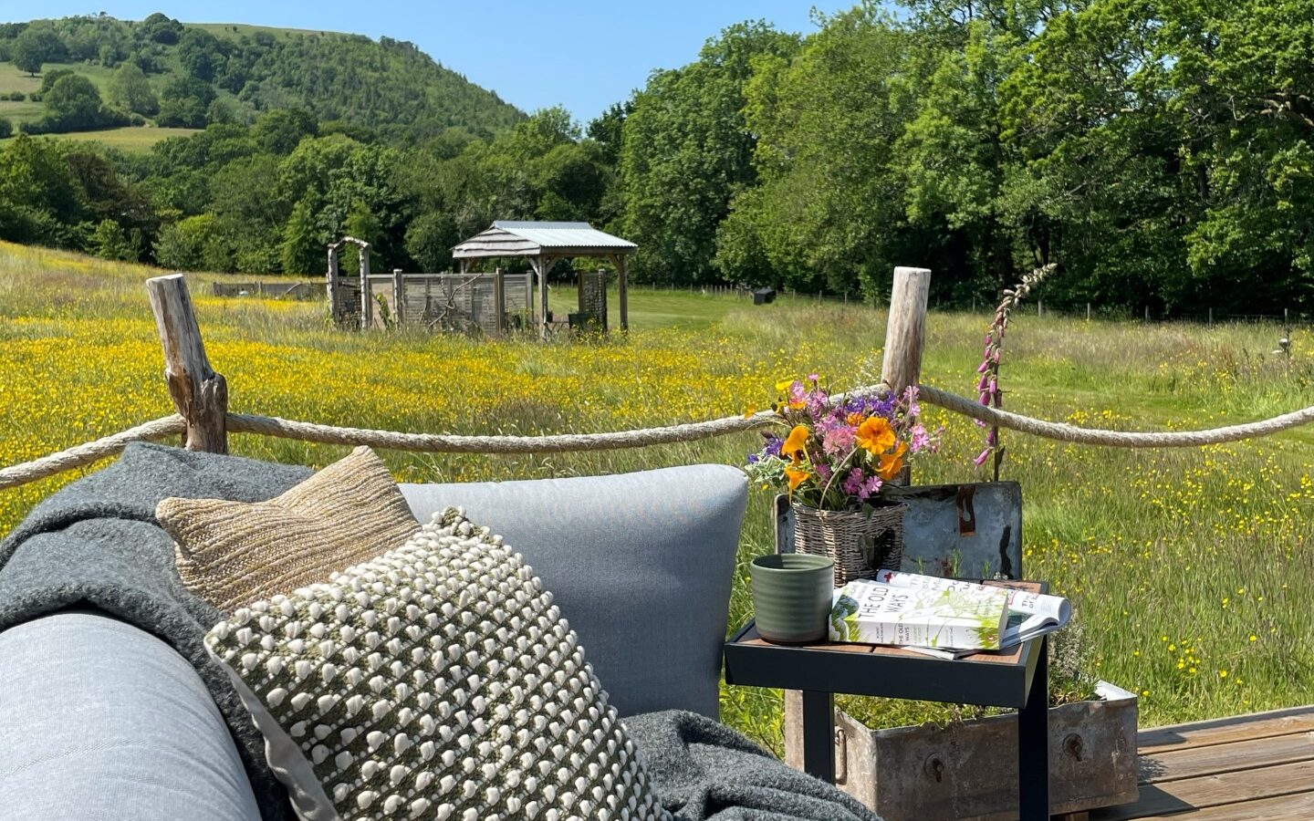 A cozy outdoor seating area with gray cushions and a textured pillow overlooks a picturesque field with wildflowers and distant hills. A small side table, branded Erwain Escapes, holds a vase of flowers, a book, and a green cup, while a rustic cabin structure stands in the background.