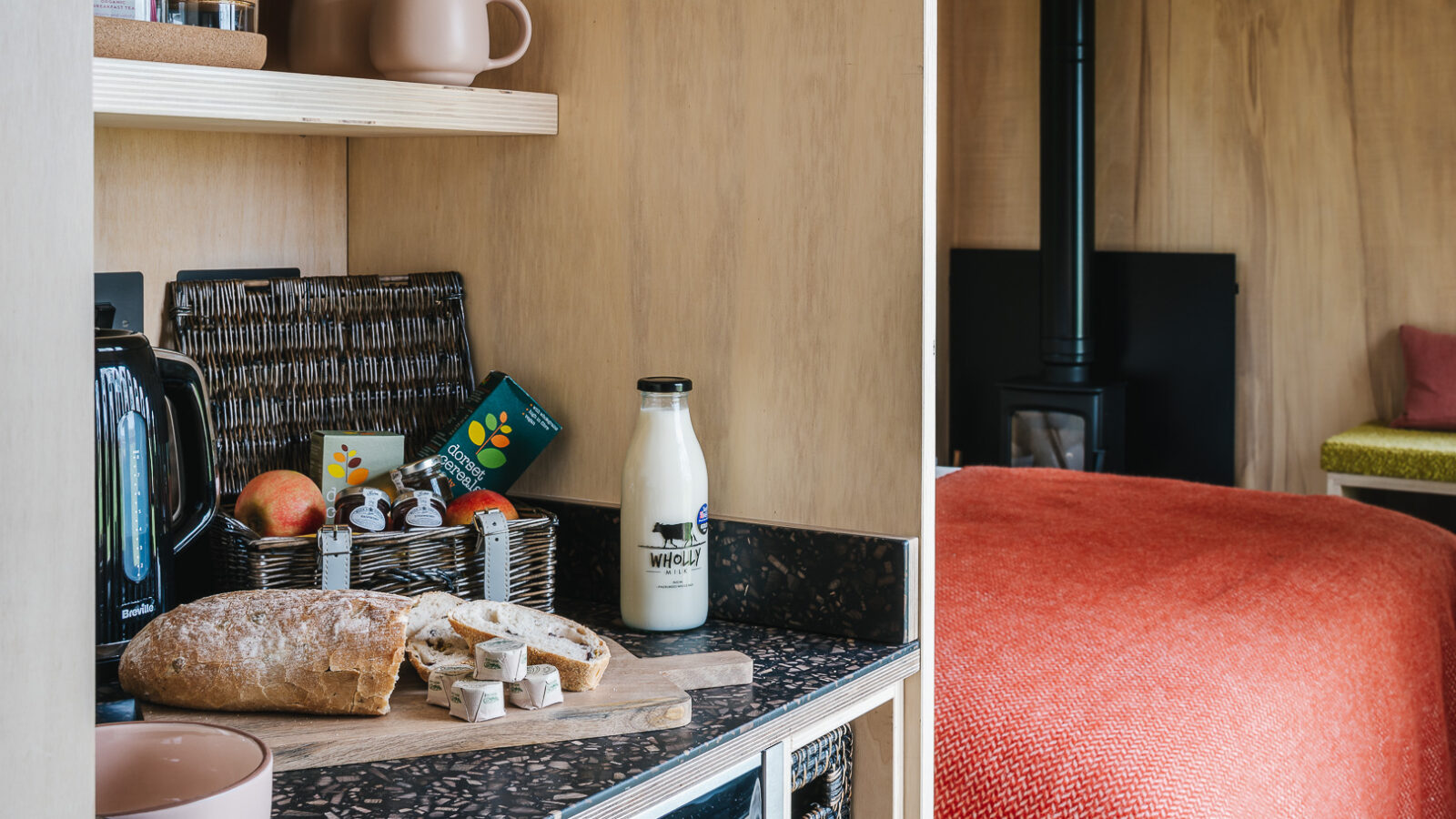 A cozy kitchen nook featuring a rustic loaf of bread on a cutting board, small packets of butter, a bottle of milk, a basket with apples and snacks, and various kitchen items. To the right, a glimpse of a bed with an orange blanket rewilds the space against the backdrop of a wood-burning stove.