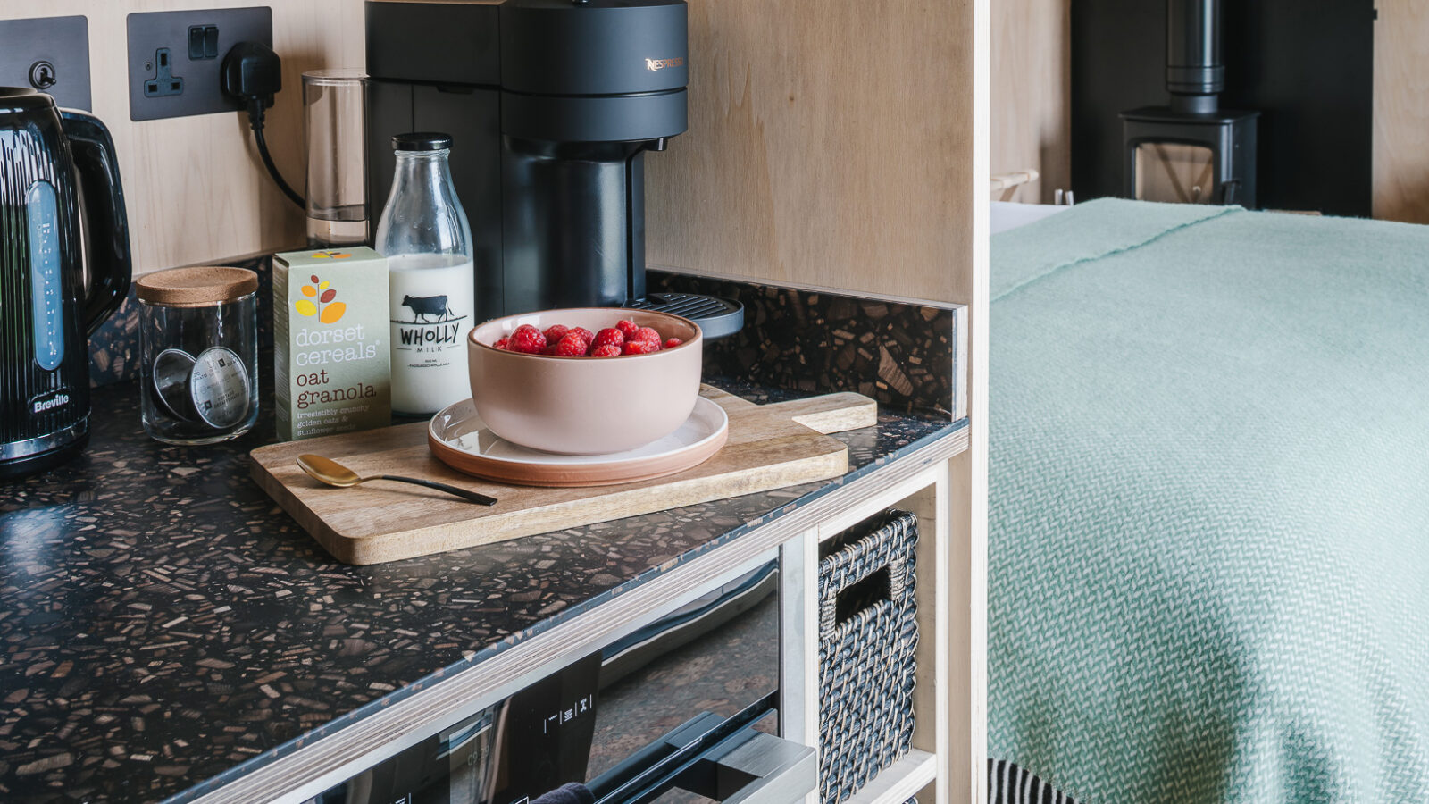 A modern cozy space featuring a kitchen counter with a black kettle, coffee machine, milk bottle, orange granola cereal box, and a bowl of cereal with strawberries on a wooden cutting board. Adjacent to it is a neatly made bed with a light green blanket that evokes the tranquility of treehouses.