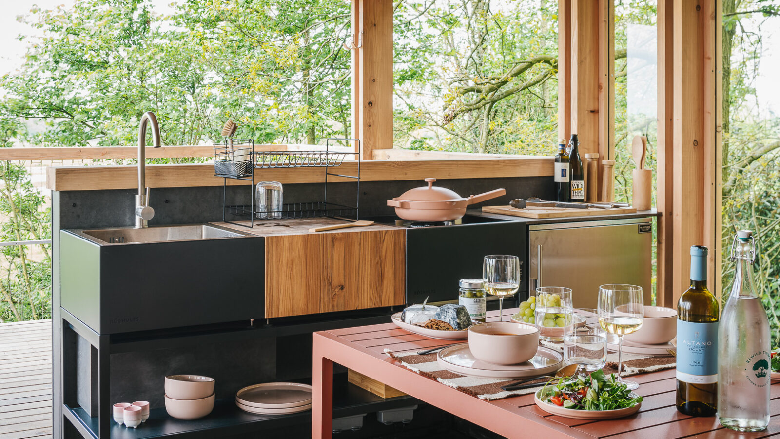 An outdoor kitchen with wooden and steel elements, featuring a modern sink, stovetop, and a small refrigerator. A table in the foreground is set with plates of salad, glasses of white wine, and a wine bottle. The setting evokes the charm of treehouses, with trees visible through open walls.