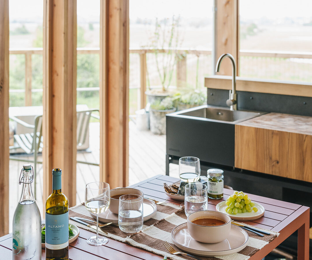 A cozy dining area in a wooden cabin features a set table with dishes, wine, and water bottles. A salad, bread, and fruit accompany a bowl of soup on each plate. Large windows showcasing a scenic outdoor view of treehouses rewild things, while a modern sink enhances the space.