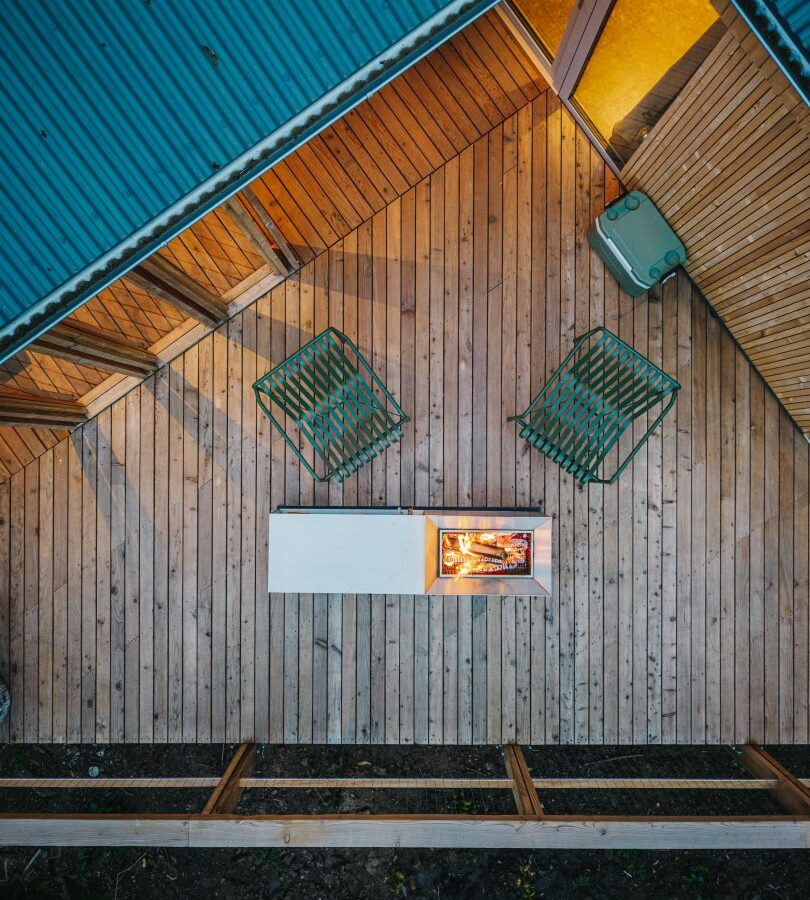 Aerial view of a wooden deck with a minimalist setup. The deck includes two green wire chairs, a modern fire pit with a small flame, and a green refrigerator against the side wall. A portion of the triangular, blue metal roof is visible, evoking the charm and tranquility reminiscent of treehouses.