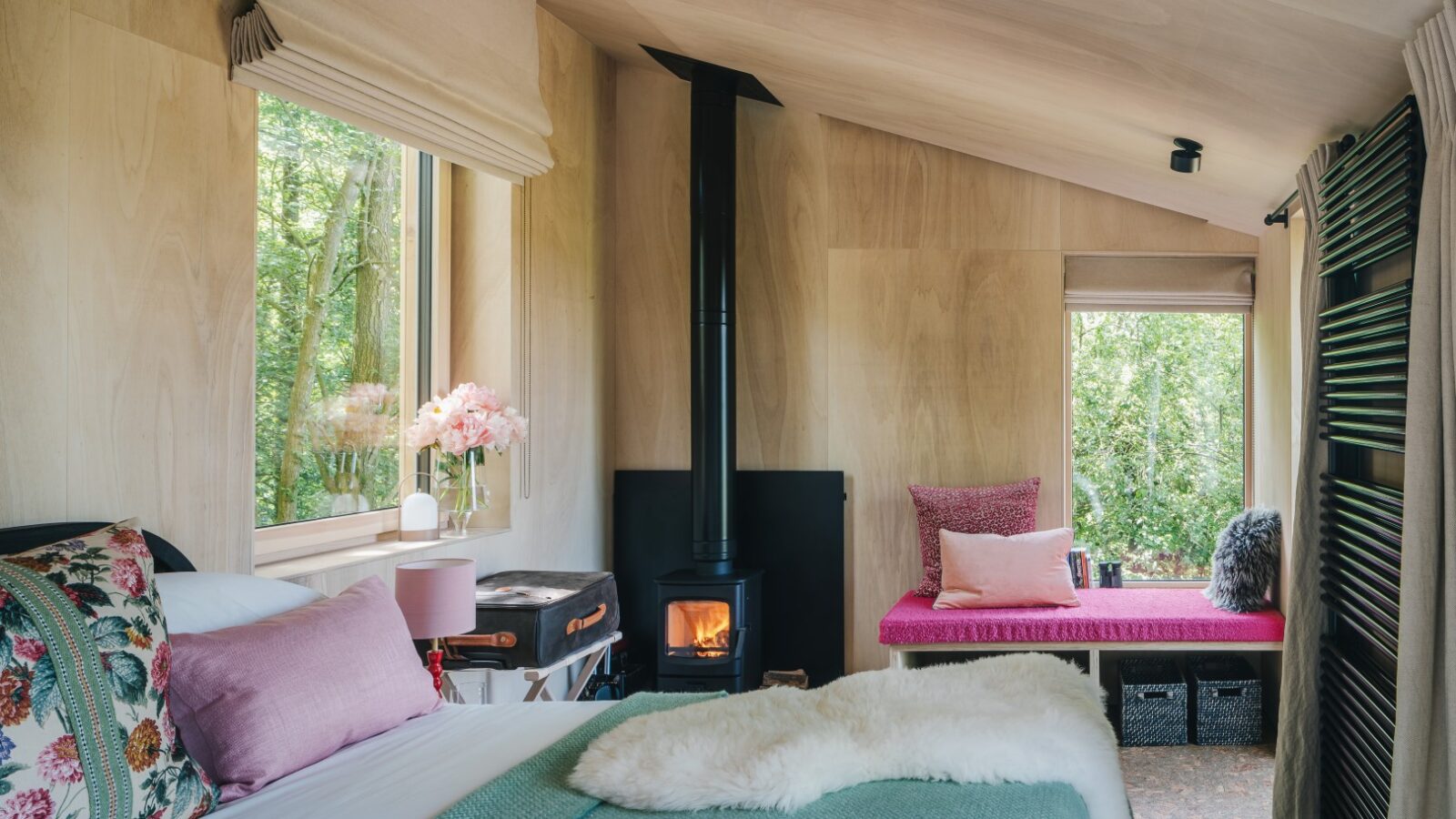 A cozy cabin interior featuring a bed with floral and pastel-colored pillows and blankets. Beside the bed, in true treehouse charm, is a small wood-burning stove with a fire burning. Two large windows allow natural light to enter, and a cushioned bench by the window adorned with pillows invites you to rewild things.