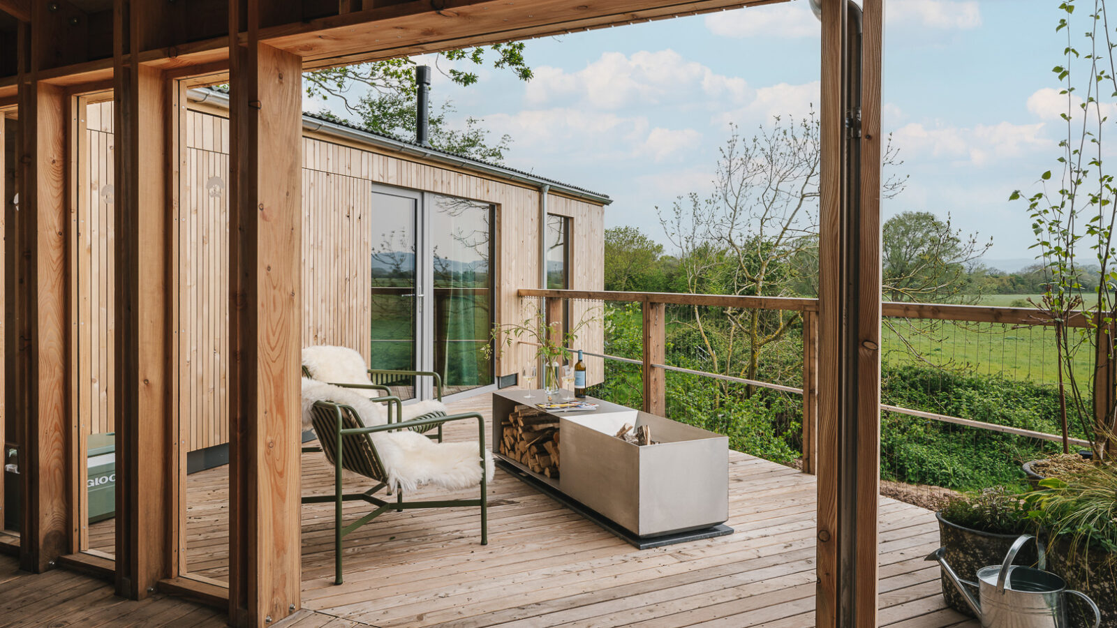 A modern wooden deck with a cozy seating area featuring two green chairs with white cushions. A rectangular fire pit topped with logs is set in the center. The background shows lush greenery and treehouses under a partly cloudy sky. A watering can is placed nearby.