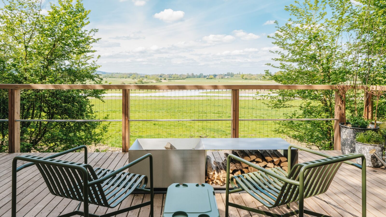 A wooden deck with two green metal chairs facing a rectangular metal fire pit and a green cooler. The deck overlooks a grassy field with trees, conjuring the feel of Treehouses, and a partly cloudy sky above. Firewood is stored beneath the fire pit, perfect for those who love to Rewild Things.