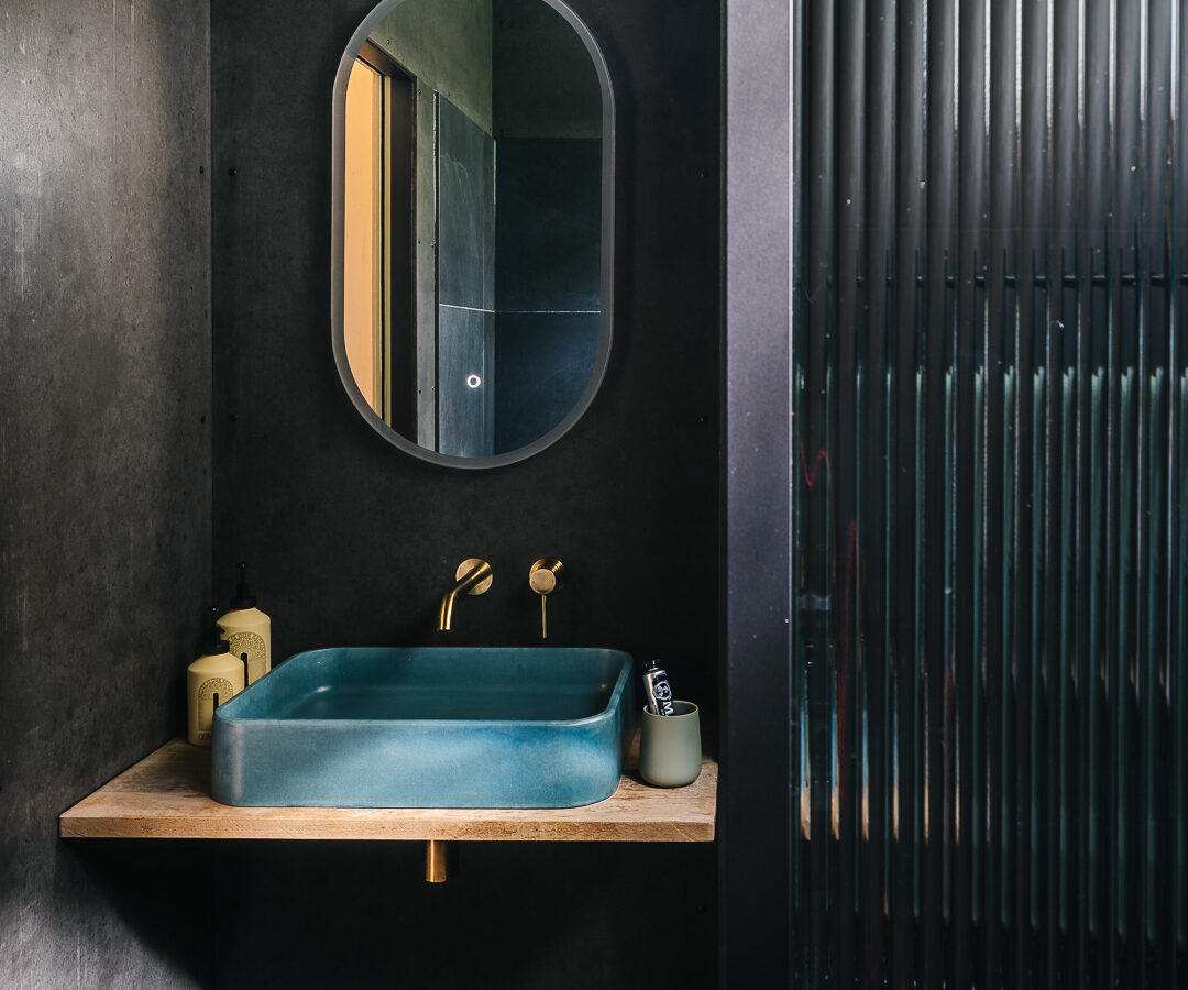 A modern, minimalist bathroom featuring a dark-toned interior and subtle nods to Rewild Things. There is a blue rectangular sink with a gold faucet, set on a wooden shelf reminiscent of treehouses. Above it, a rounded rectangular mirror is mounted on the wall. A small wooden stool is placed underneath the sink.