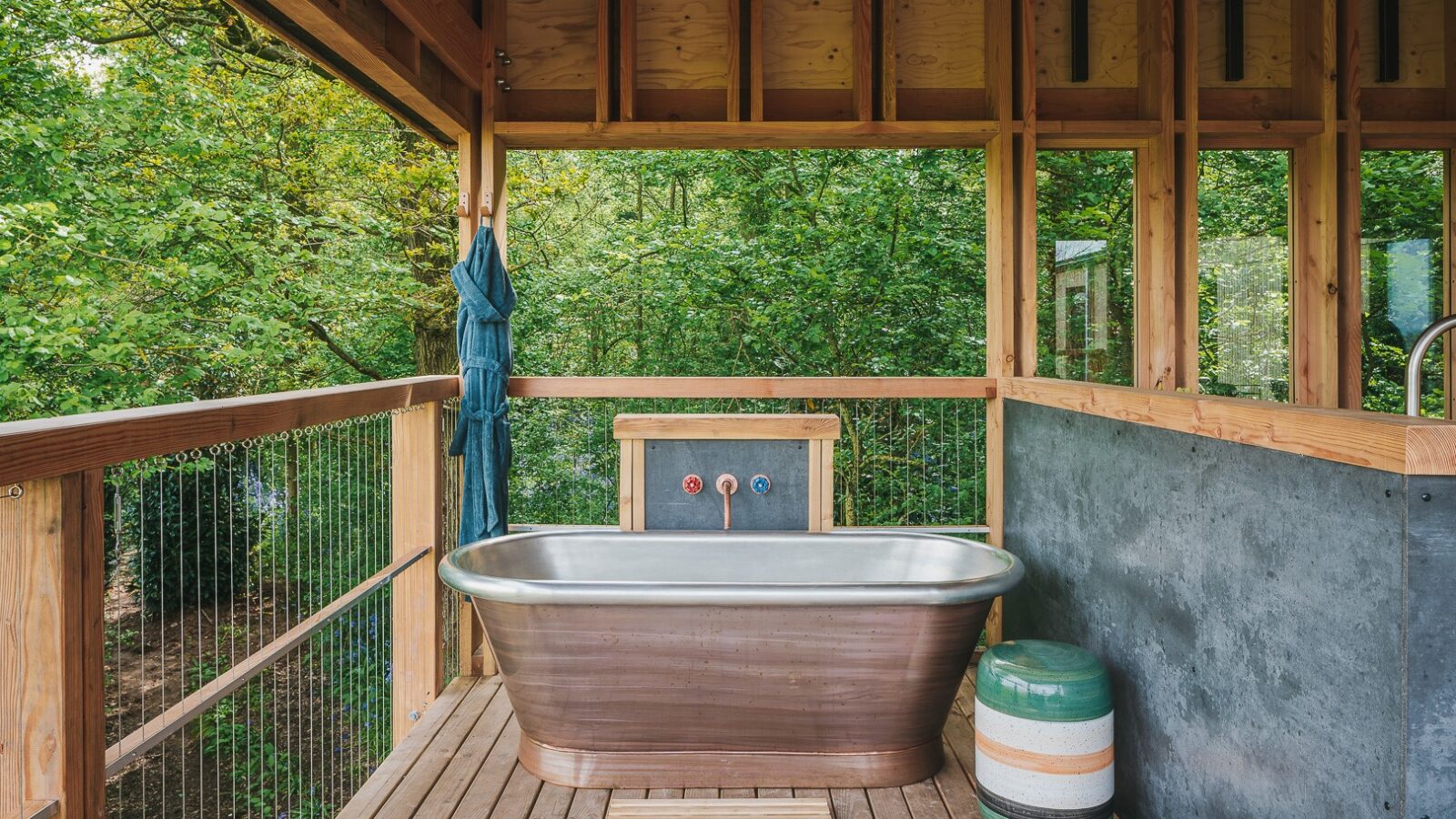 A rustic outdoor bathroom with a wooden deck features a large, metal bathtub with wooden accents. The background includes a railing overlooking lush green trees, reminiscent of treehouses. There is a small barrel with striped colors beside the bathtub and towels hanging nearby, perfect for rewilding things.