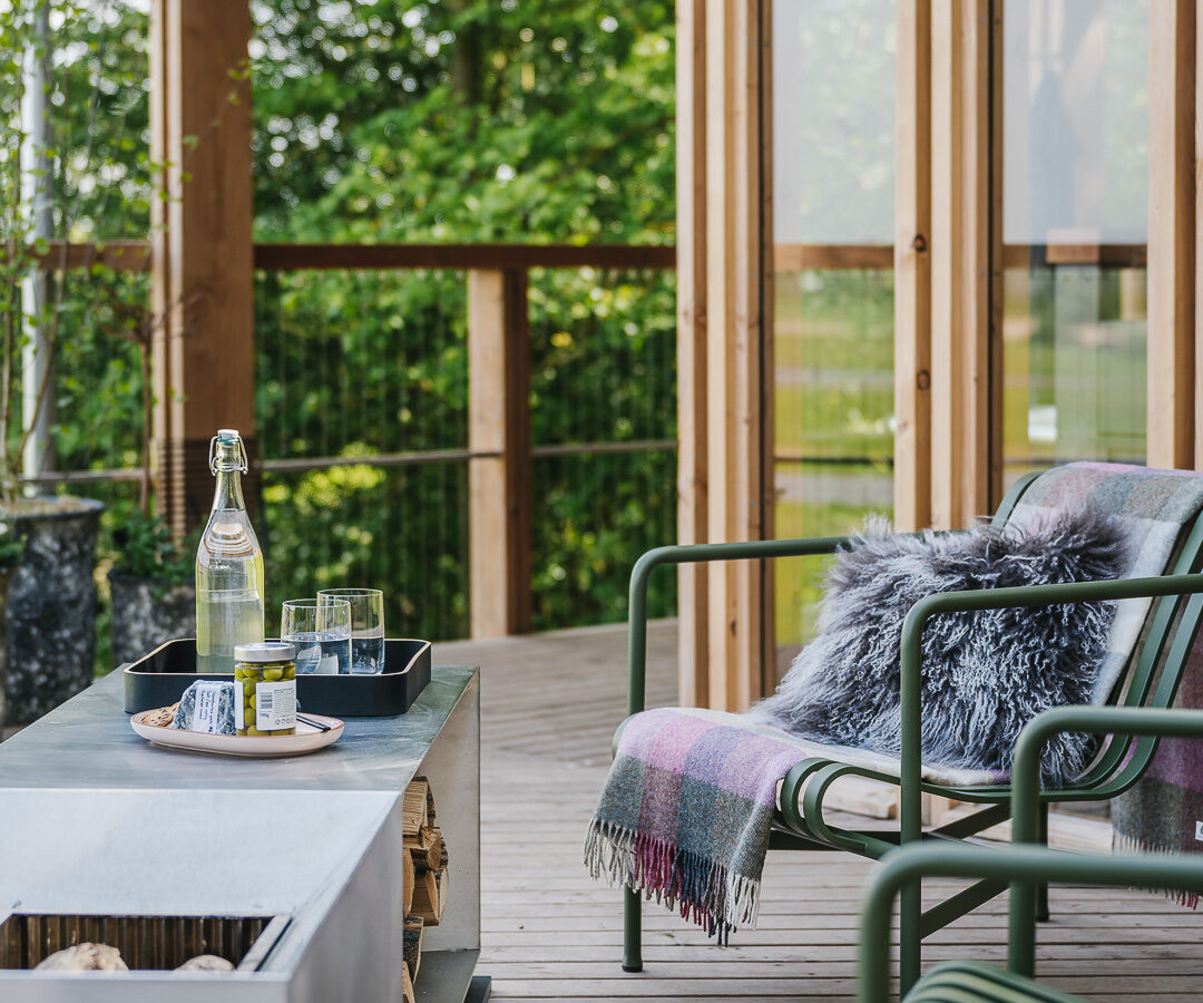 A cozy outdoor patio with modern wooden architecture, featuring a green chair draped with a colorful blanket and fur pillow. There's a sleek table with a bottle of water, glasses, and a small plant. The ambiance evokes the charm of treehouses, with large glass doors and lush greenery in the background.