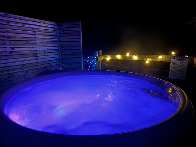 At Rowan Tree Retreat, a circular hot tub illuminated with blue and purple lights is surrounded by wooden paneling on one side and a boundary adorned with string lights on the other. A safety notice is posted nearby under the nighttime sky.