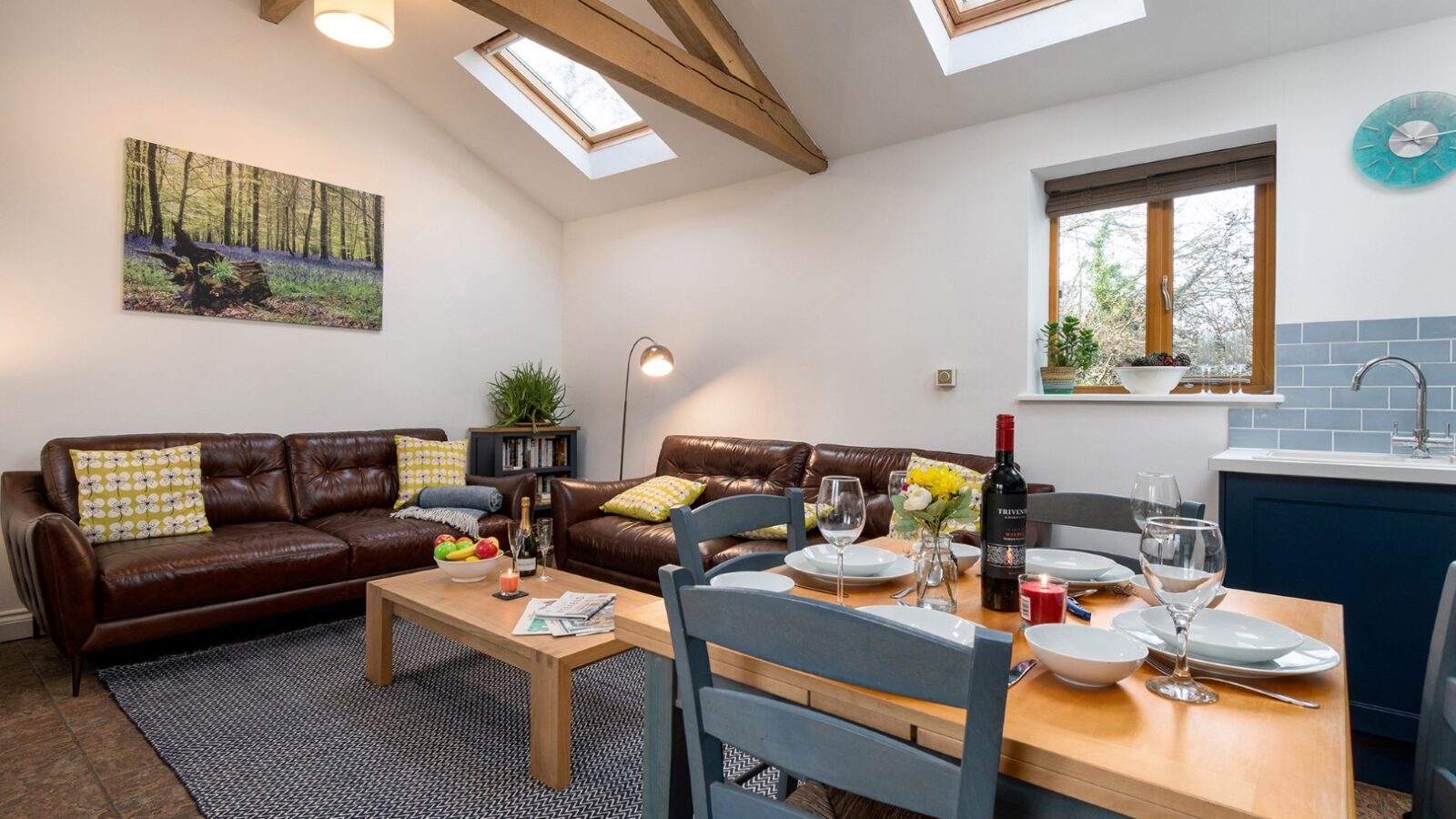 A cozy living-dining area at the Rowan Tree Retreat features a brown leather sofa set, a wooden coffee table with assorted fruits, and a set dining table with plates and wine. The room has skylights, exposed wooden beams, and a serene forest painting on the wall. A kitchen counter is in the background.