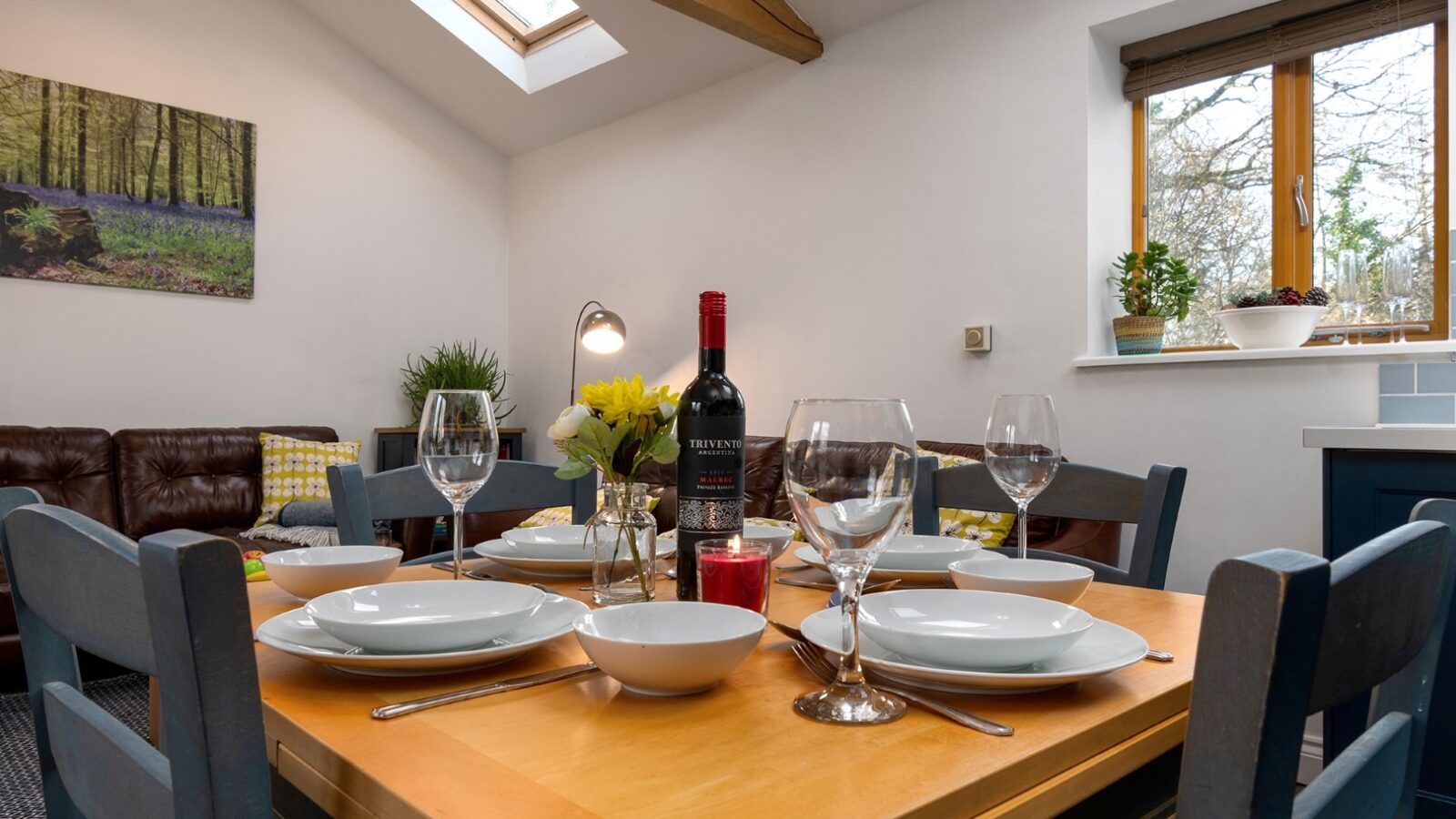 A cozy dining area at Rowan Tree Retreat features a wooden table set for four, adorned with white plates, bowls, wine glasses, and a bottle of red wine. The room boasts a skylight, plants on the windowsill, and forest artwork on the wall. A couch with cushions is visible in the background.