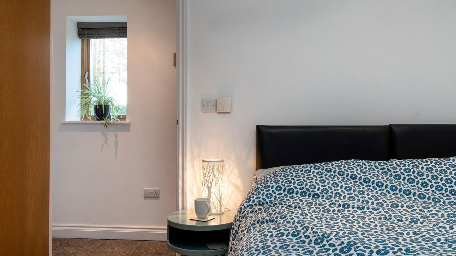 A cozy bedroom at Rowan Tree Retreat features a bed covered in a blue and white patterned duvet. Beside the bed is a small round table with a modern lamp and a cup. A window, with a plant on the sill, brings in natural light, and dark-colored flooring completes this serene escape.