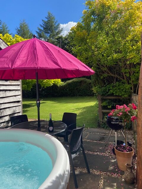 A cozy outdoor patio with a round hot tub next to a small table and chairs shaded by a bright pink umbrella. A bottle of wine rests on the table. Nearby, a barbecue grill and potted flowering plants are visible, surrounded by lush greenery—a true Tree Retreat.