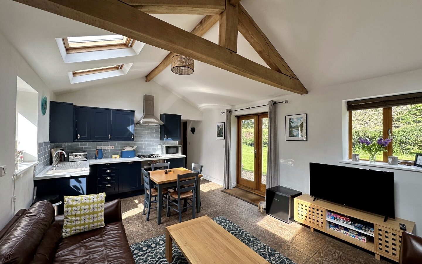 A cozy open-plan living space at Rowan Tree Retreat with a brown leather sofa, wooden coffee table, and flat-screen TV on a stand. The kitchen area features blue cabinets, a stove, and a dining table with chairs. The room is bright with skylights, a large window, and glass doors leading outside.