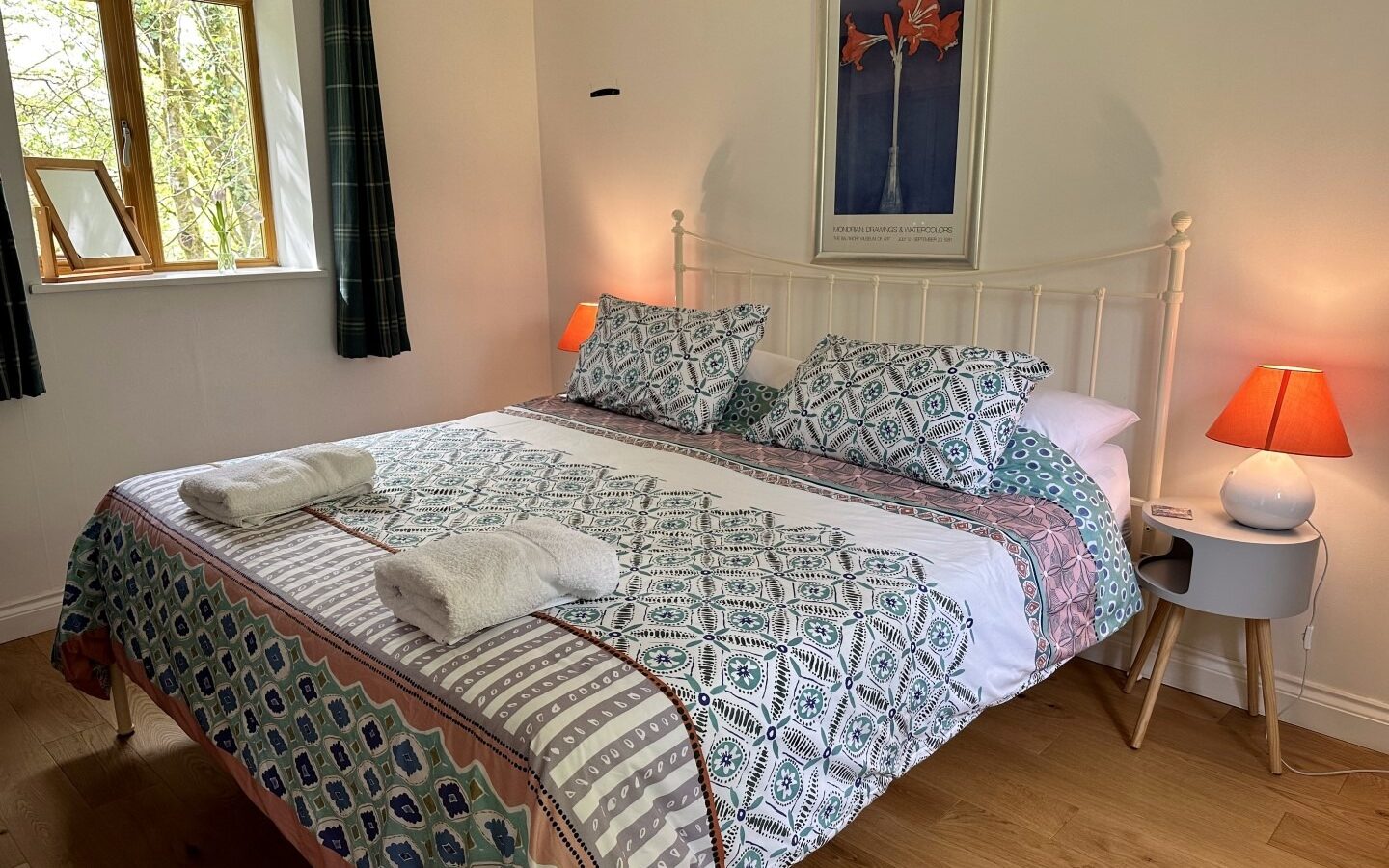 A cozy bedroom at Rowan Tree Retreat features a neatly made bed with multi-patterned bedding, two white pillows, and two folded towels at the foot. Beside the bed are two small nightstands with orange-shaded lamps. A window with dark curtains brings in natural light, and a framed picture hangs above the bed.