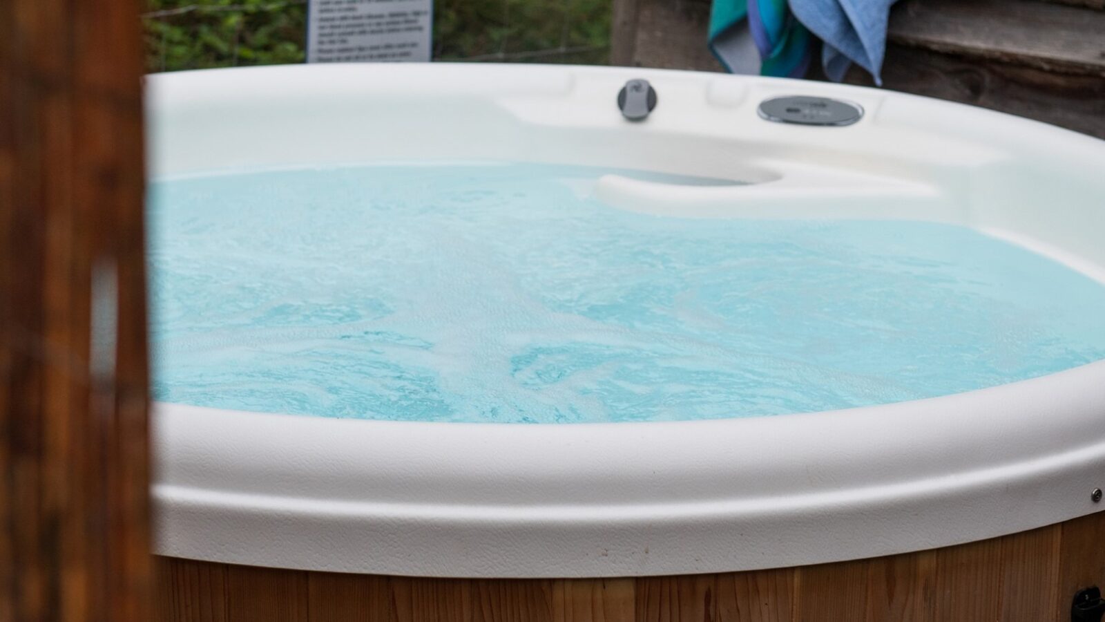 A close-up view of a filled hot tub outdoors at Rowan Tree Retreat. The hot tub is circular with light blue, bubbling water. A towel is draped over a railing in the background, and a sign with text is slightly visible. Some greenery is seen beyond the hot tub.