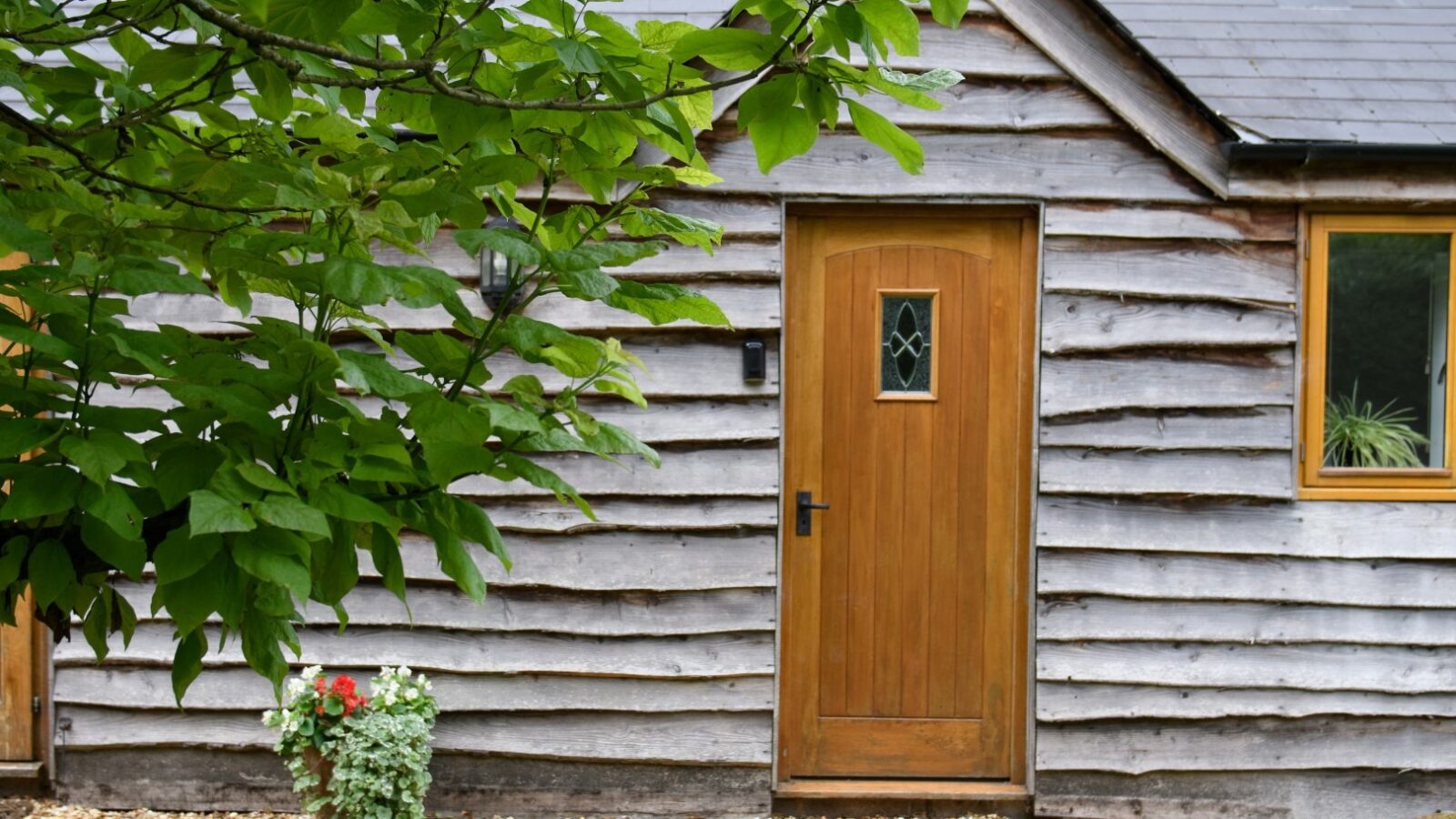 A small cottage retreat features wooden paneling, a wooden front door with a glass pane, and a window to the right. A leafy Rowan tree branch partially obscures the scene. There is a potted plant with red and white flowers by the entrance. The ground is covered with pebbles.