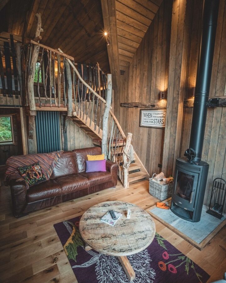 A cozy wooden cabin interior at Baxby Manor featuring a rustic staircase, a leather sofa adorned with colorful cushions and a blanket, a circular rustic coffee table, and a wood-burning stove. A sign on the wall reads, 