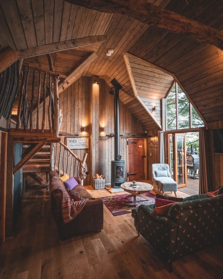 Cozy cabin interior with wooden walls and ceiling, featuring a staircase, a wood-burning stove, a couch, armchairs, and a round coffee table. Large windows and sliding doors offer a view of the trees outside at Rufus's Roost in Baxby Manor's enchanting treehouse, creating a warm and rustic atmosphere.