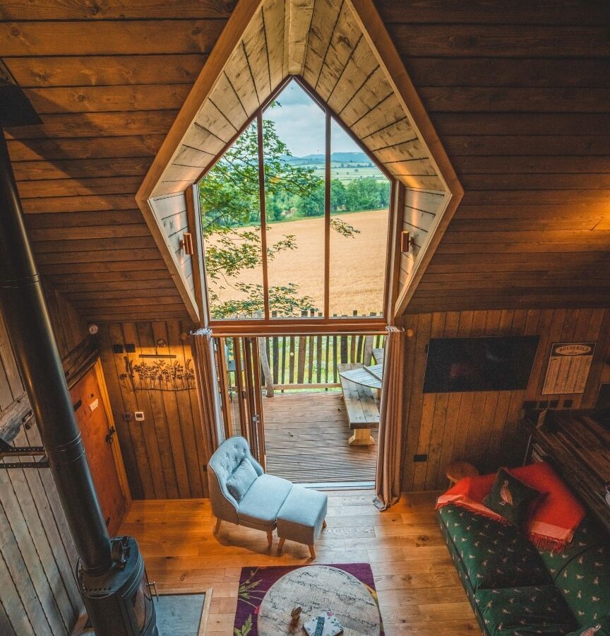 A cozy, rustic interior of Rufus's Roost Treehouse at Baxby Manor features a large, diamond-shaped window offering a view of a meadow. The room includes a wood stove, a green couch, a round coffee table, a grey armchair, and a balcony with a bench. The cabin has wooden walls and flooring.