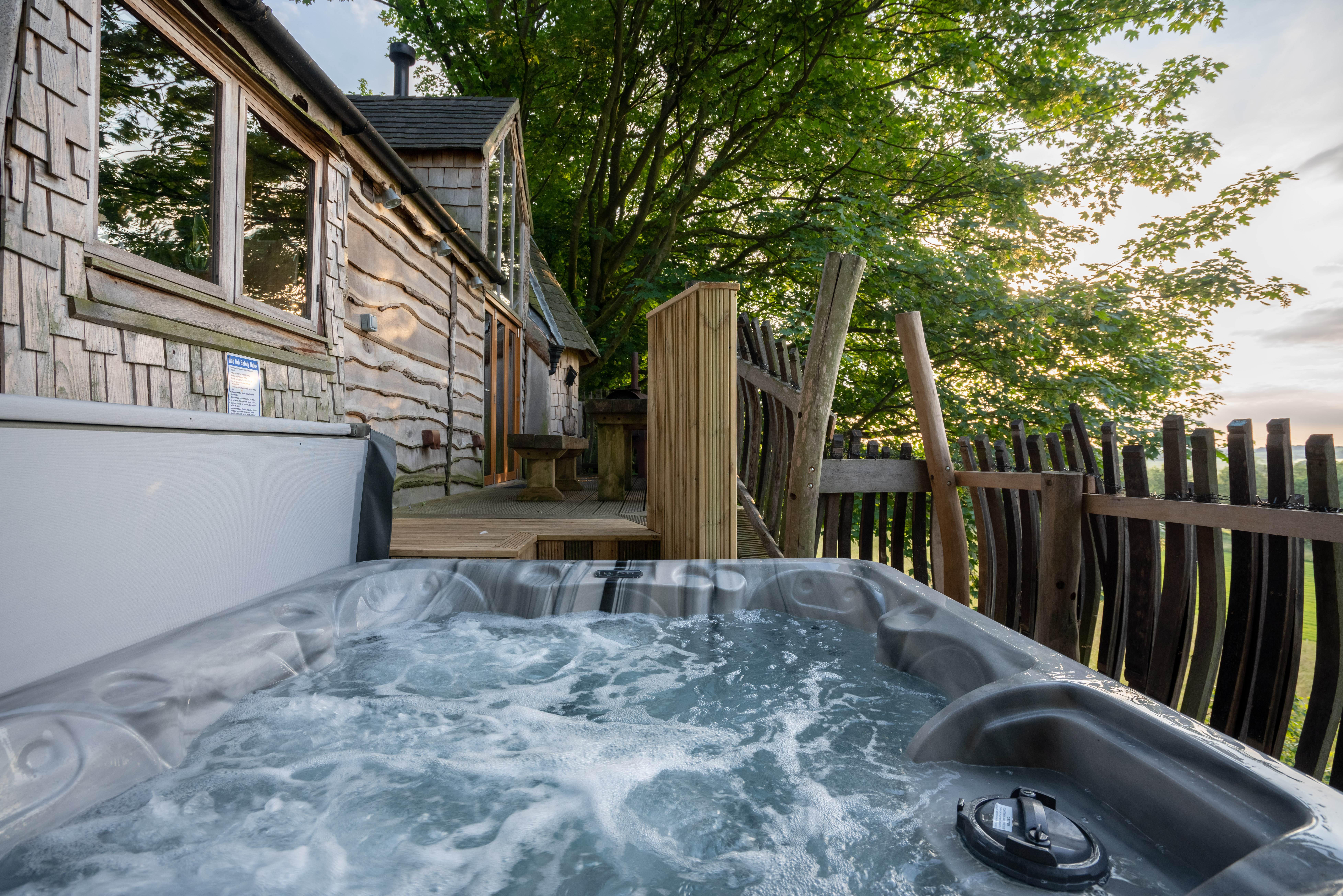 A bubbling hot tub is positioned on a wooden deck in front of a rustic cabin, reminiscent of Rufus's Roost Treehouse at Baxby Manor. The cabin features a mix of wood and stone exterior, large windows, and a small porch. Trees and a wooden fence surround the area, with the sun setting in the distance.