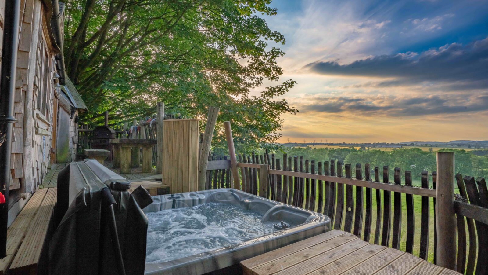 A serene outdoor scene featuring a hot tub on a wooden deck next to Rufus's Roost, a rustic treehouse cabin. The deck is bordered by a wooden fence, overlooking a lush green landscape and a distant horizon under a partly cloudy sky during sunset.