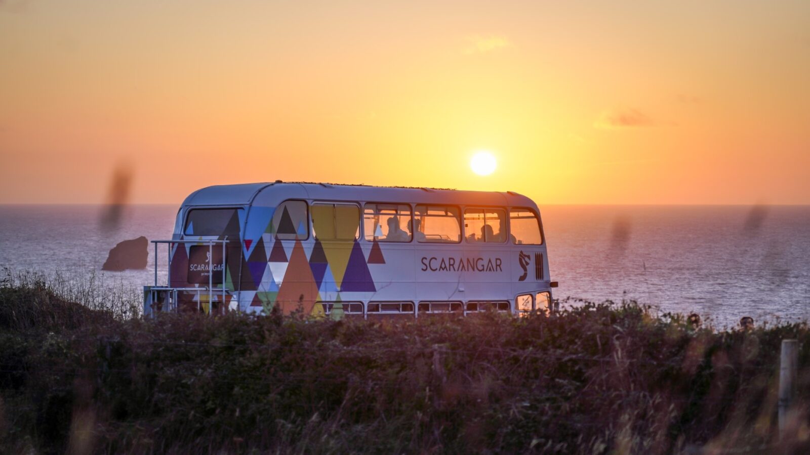 A brightly colored bus with geometric patterns is parked on a cliffside, overlooking the ocean at sunset. The sun is low on the horizon, casting a warm glow on the water and surrounding landscape. The sky, reminiscent of Scaragnar's legendary sunsets, is a gradient of orange and yellow hues.