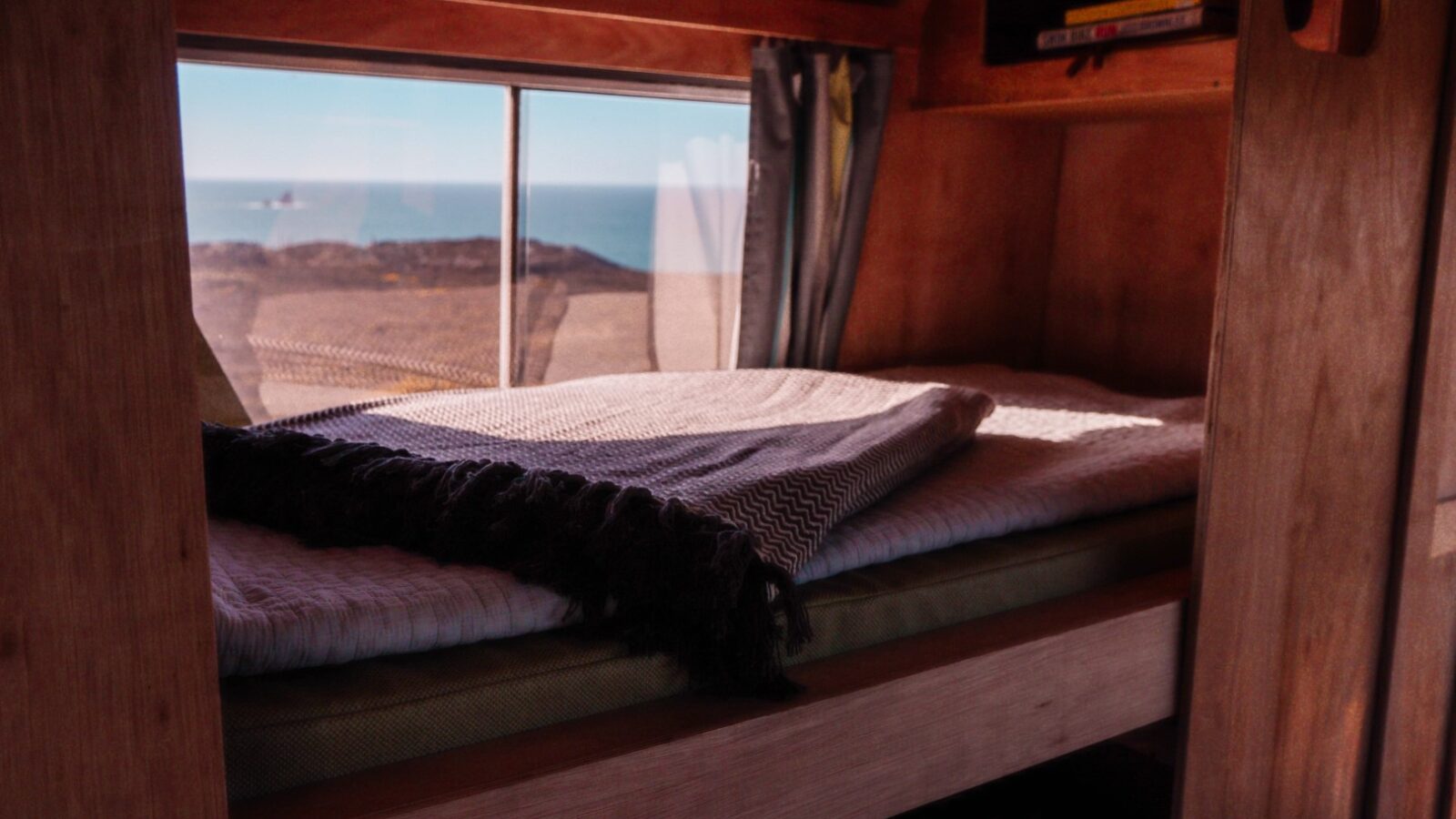 A cozy wooden bunk bed with a neatly folded gray blanket. A small window above reveals a picturesque view of the Scarangar coastal landscape with the ocean in the distance. Shelving above the bed holds a few books. The interior emits a warm, welcoming ambiance.